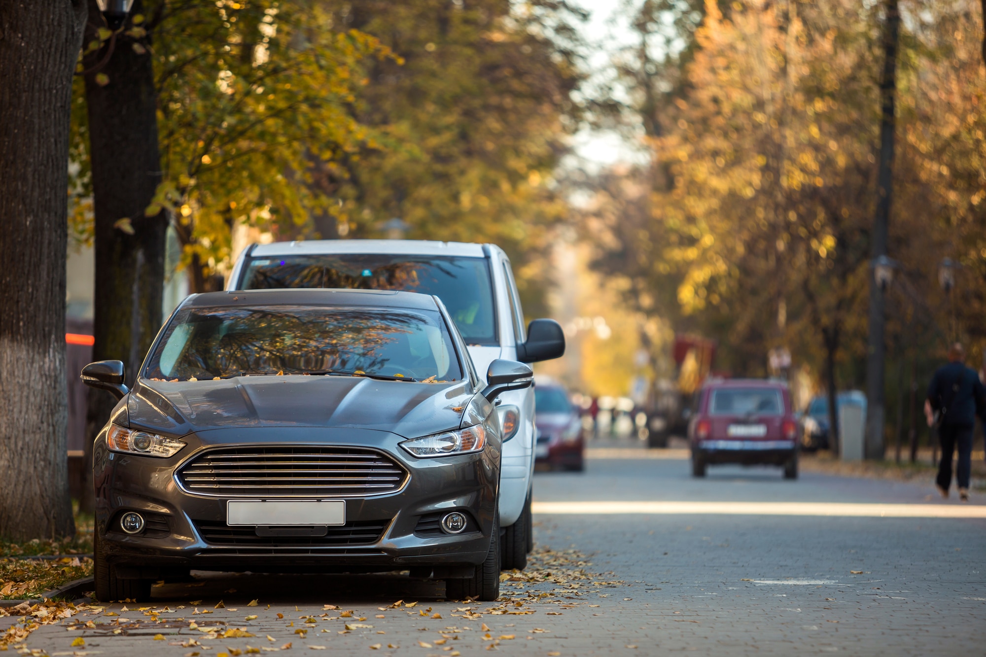 Front view of gray luxury car parked by the curb on tree-lined street on sunny autumn day w/ blurred vehicles and walking people