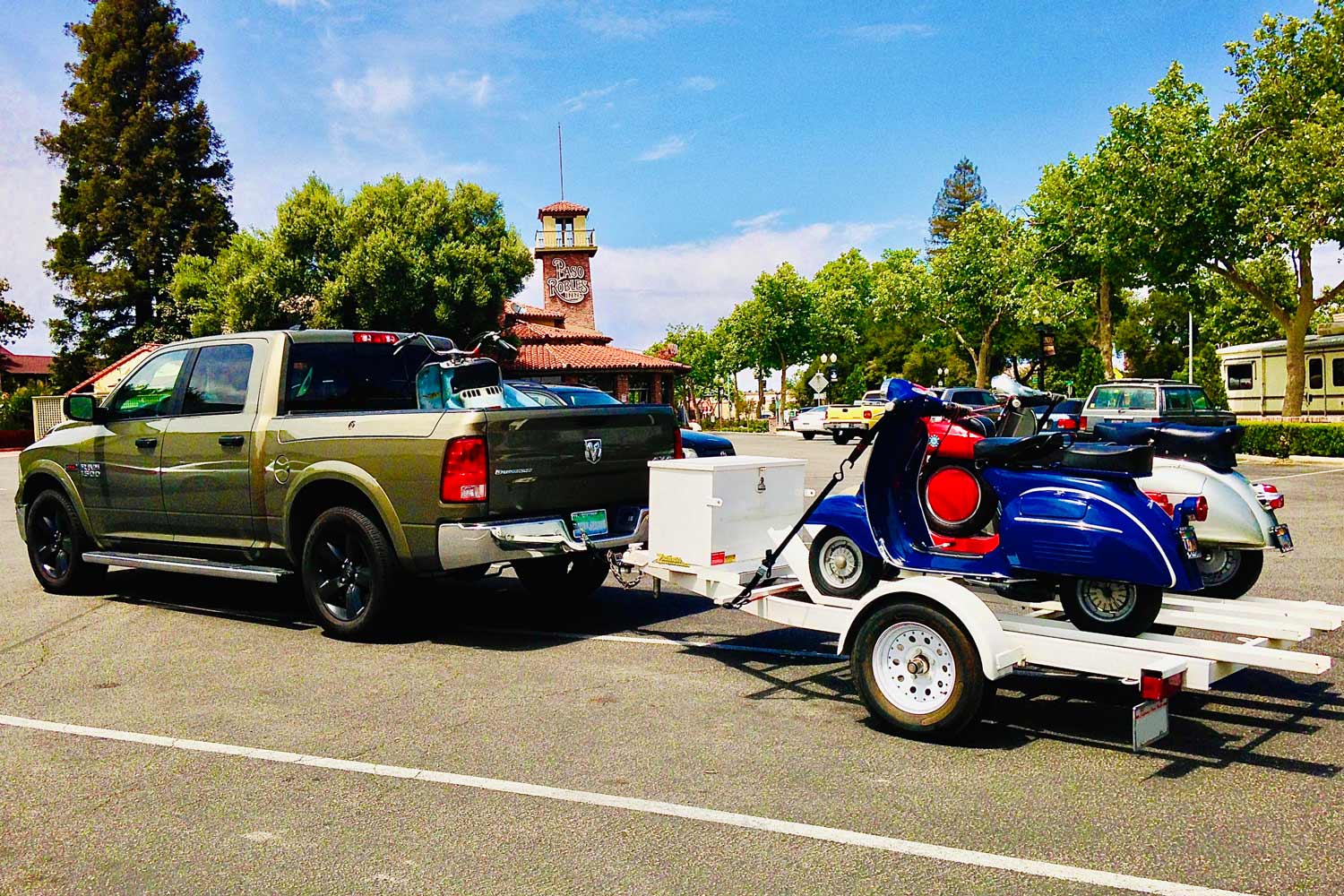 A green Ram truck tows a white motorcycle trailer with three vintage scooters riding on it.