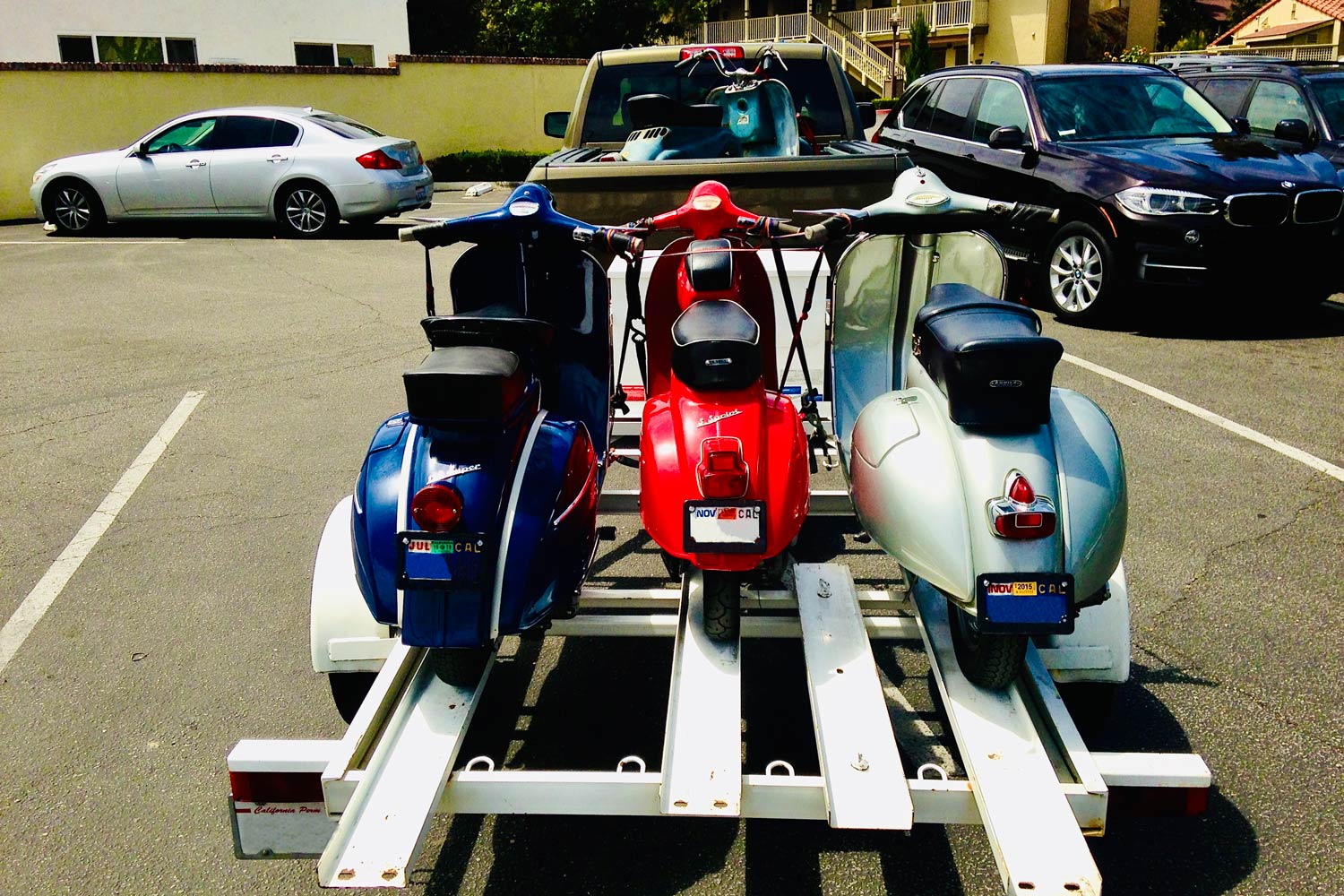 A blue, a red, and a silver vintage Vespa sit on a white motorcycle trailer with a vintage blue Henkel scooter sitting in the bed of a green Ram truck.