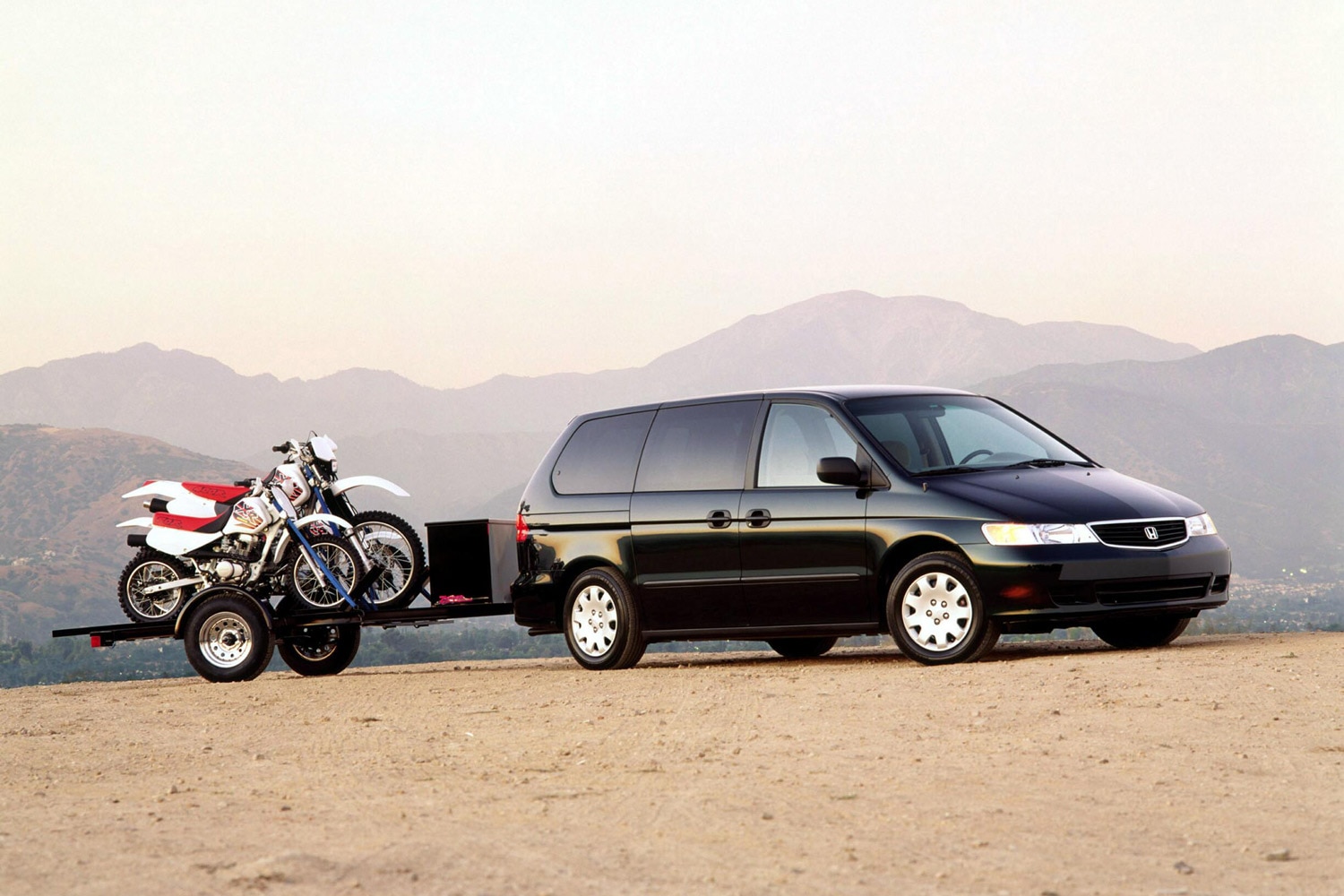 A black Honda minivan tows a black trailer holding two Honda dirt bikes.