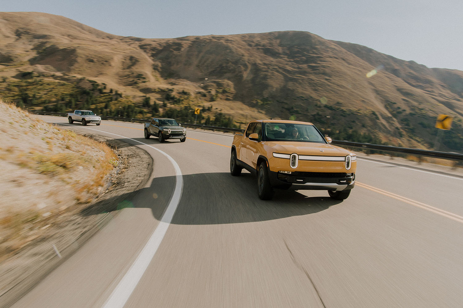 Rivian R1T in orange driving on mountain road with other trucks behind it