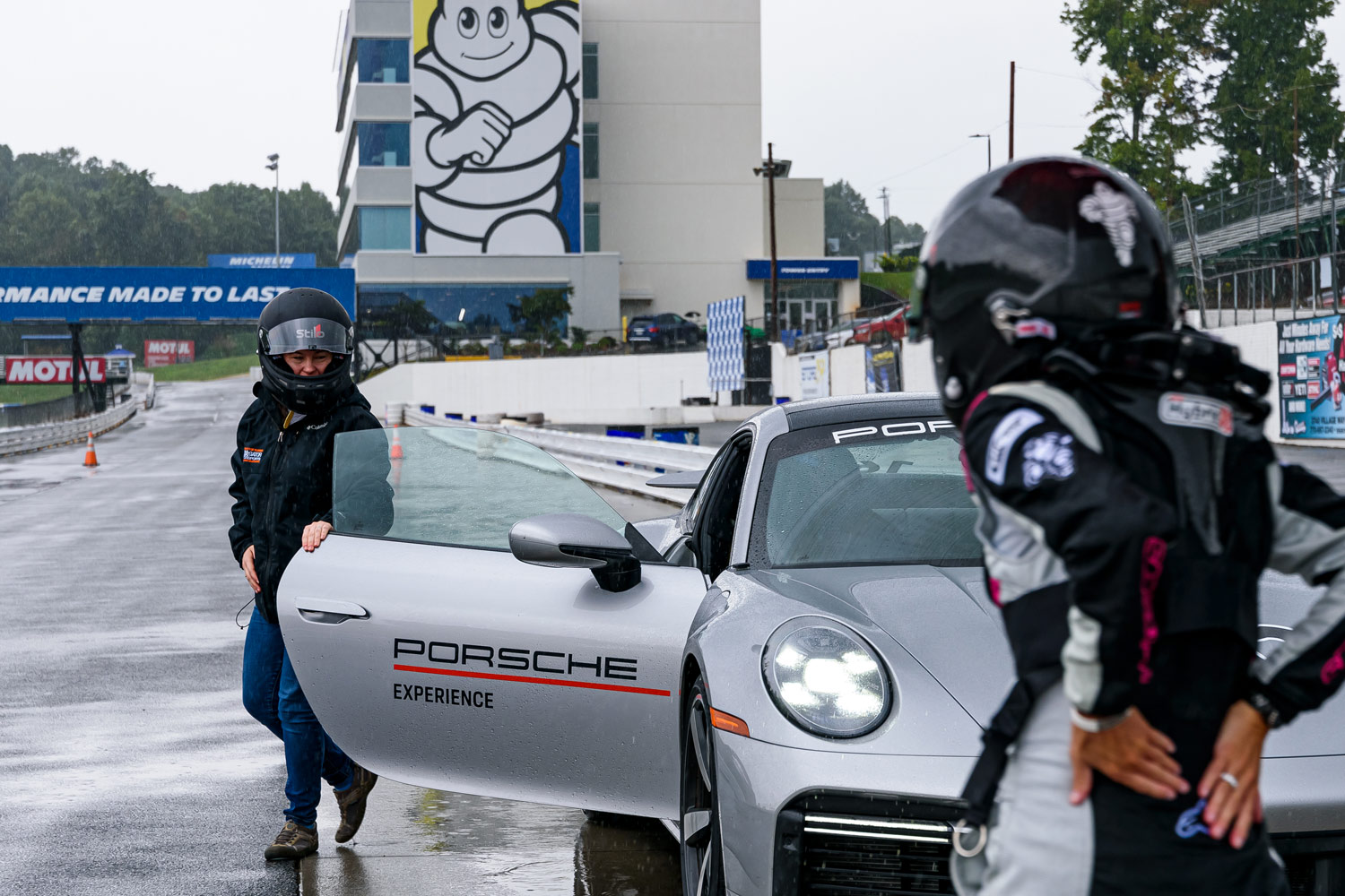 Two people wearing helmets preparing to enter a silver Porsche.