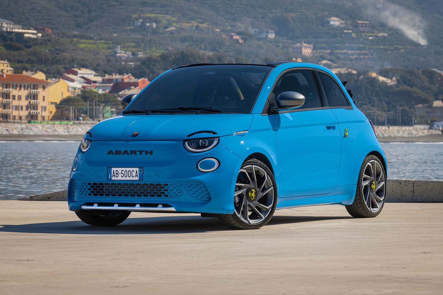 Fiat 500 in bright blue parked in front of a small body of water and a city on a hill