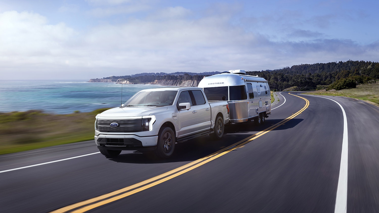 Ford F-150 Lightning EV in white tows a large Airstream camper along a coastline.