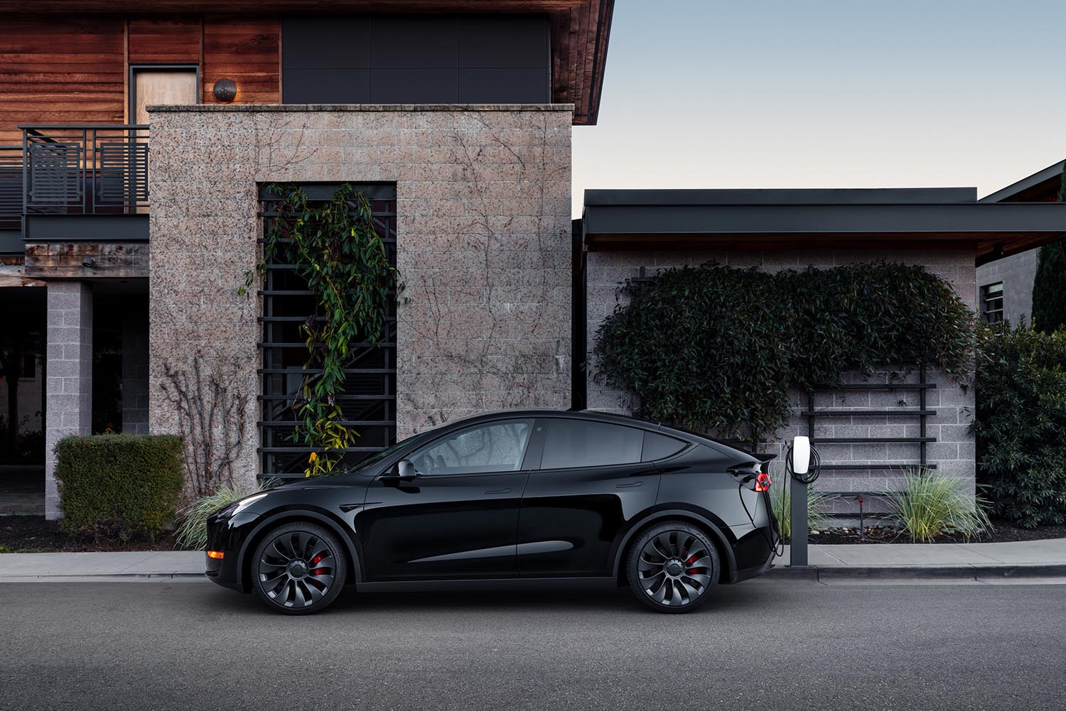 Tesla Model Y in black charges in front of a large house with a stone and wood facade.