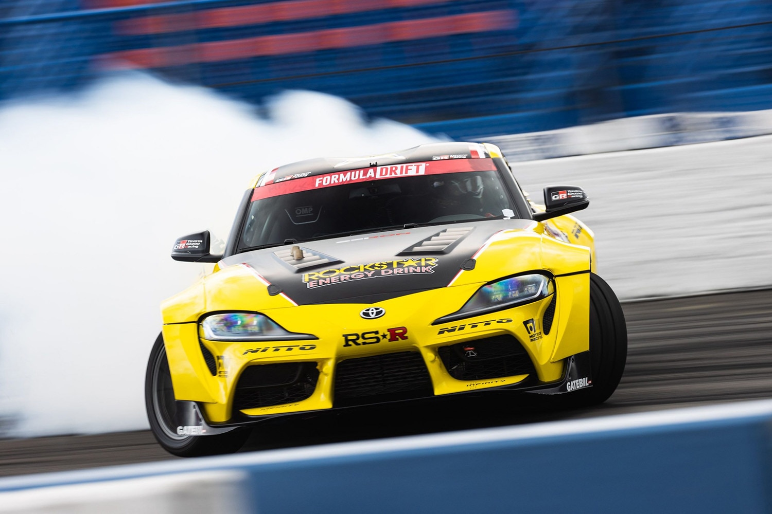 Toyota Supra in yellow race livery leaving a wake of white smoke while drifting around a track