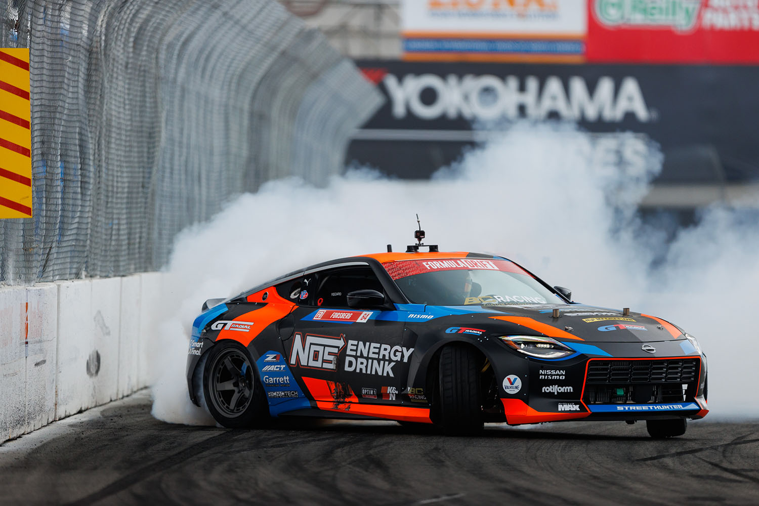 Nissan Z in black, orange, and blue with white smoke behind it drifting on a track
