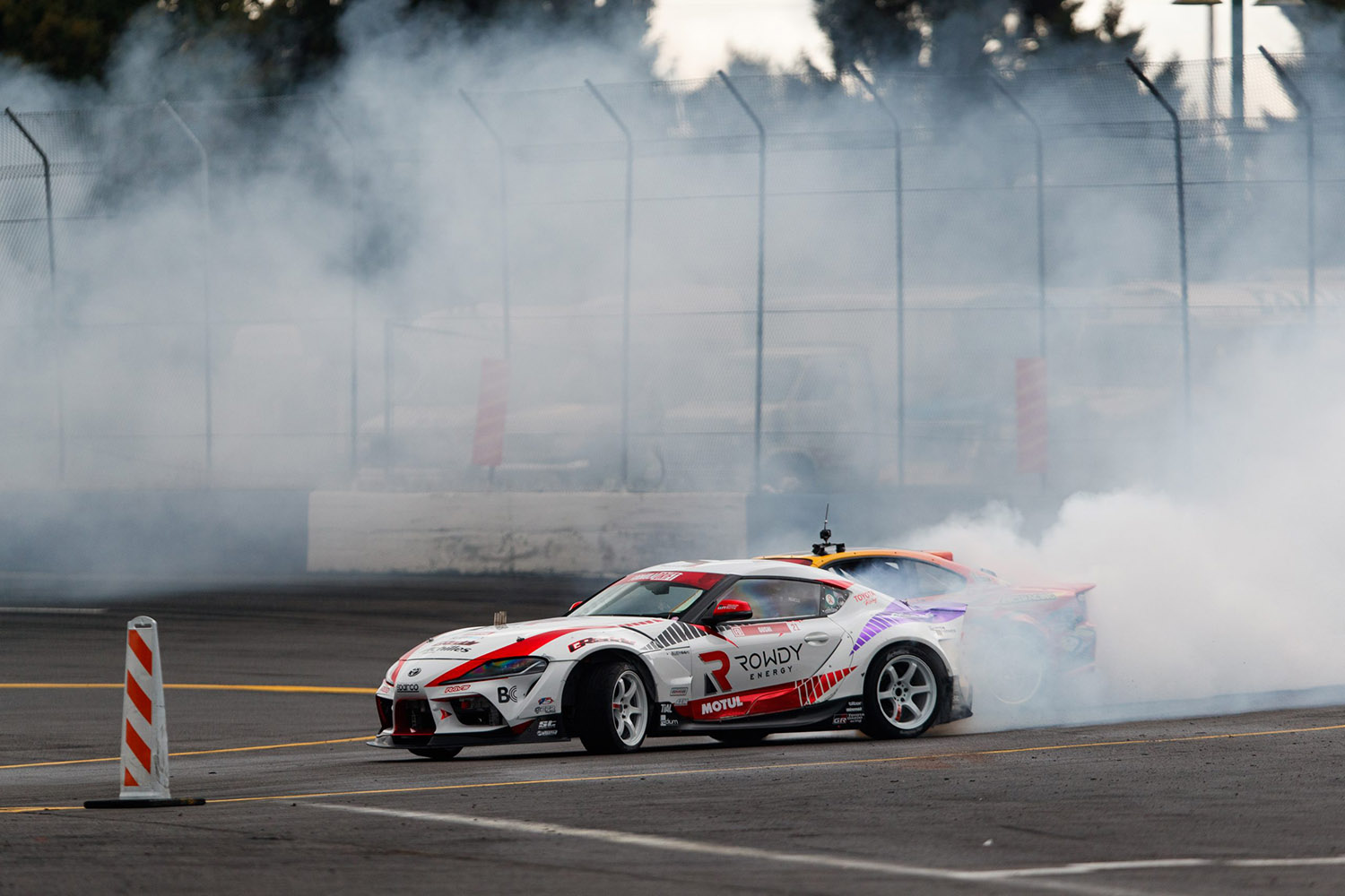 Toyota Supra in white and red drifting with white smoke and a competitor behind it on a track