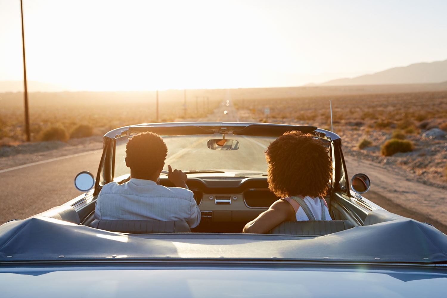 Couple driving in a blue convertible on a desert highway at sunset.