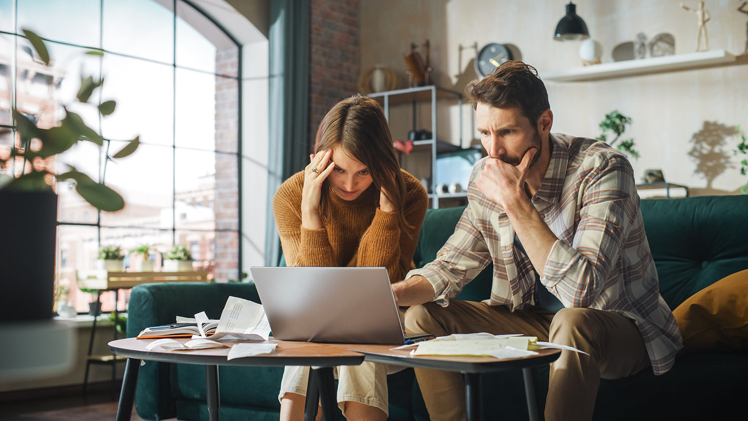 Two people sit on a green couch looking worried with a laptop and papers in front of them