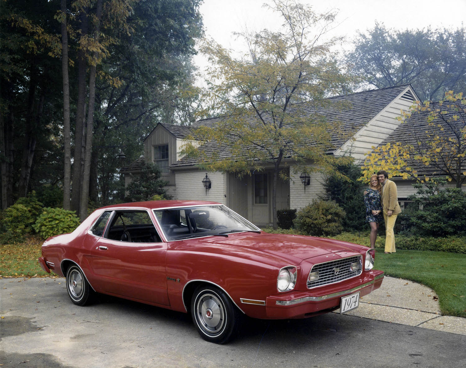A couple gazes at a red 1974 Ford Mustang II with impact bumpers parked in front of a house