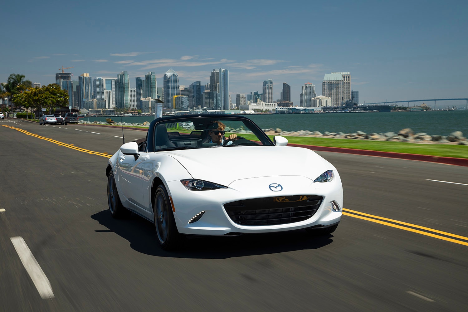 2023 Mazda MX-5 Miata in white driving along the coast with a city skyline in the background