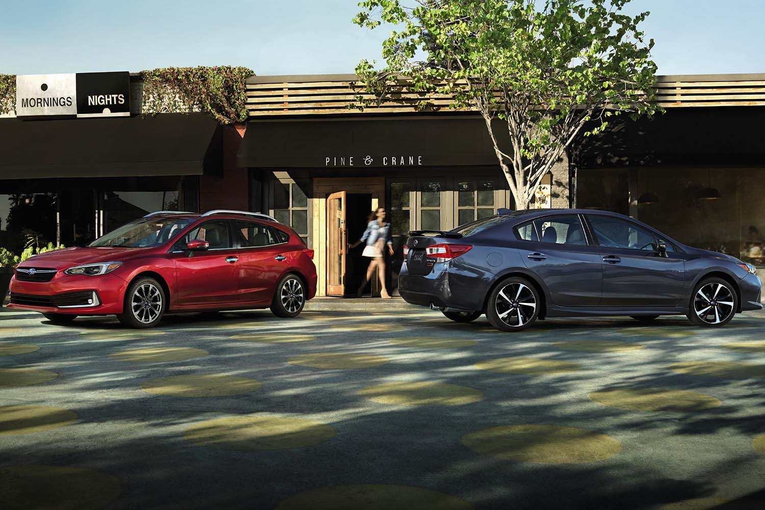 2023 Subaru Impreza 5-Door in red parked near a charcoal Impreza sedan in front of a trendy-looking coffee shop