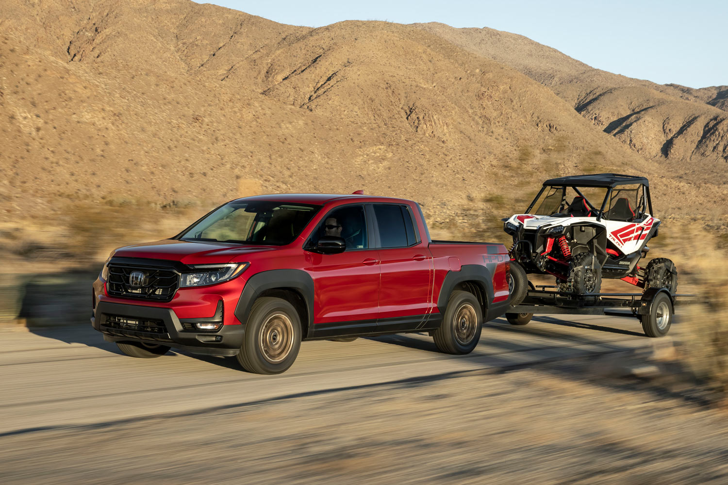 2023 Honda Ridgeline in red towing an off-road vehicle on a trailer down a dirt road