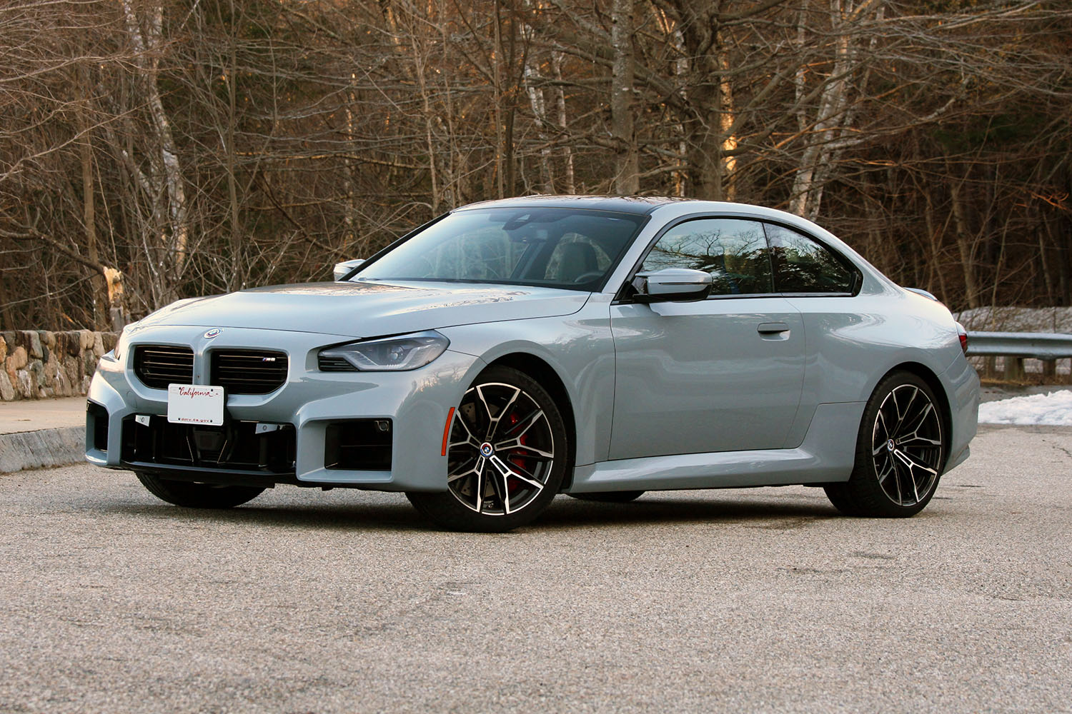 Front three-quarter shot of a 2023 BMW M2 in Brooklyn Grey Metallic parked in front of a wooded area in New England