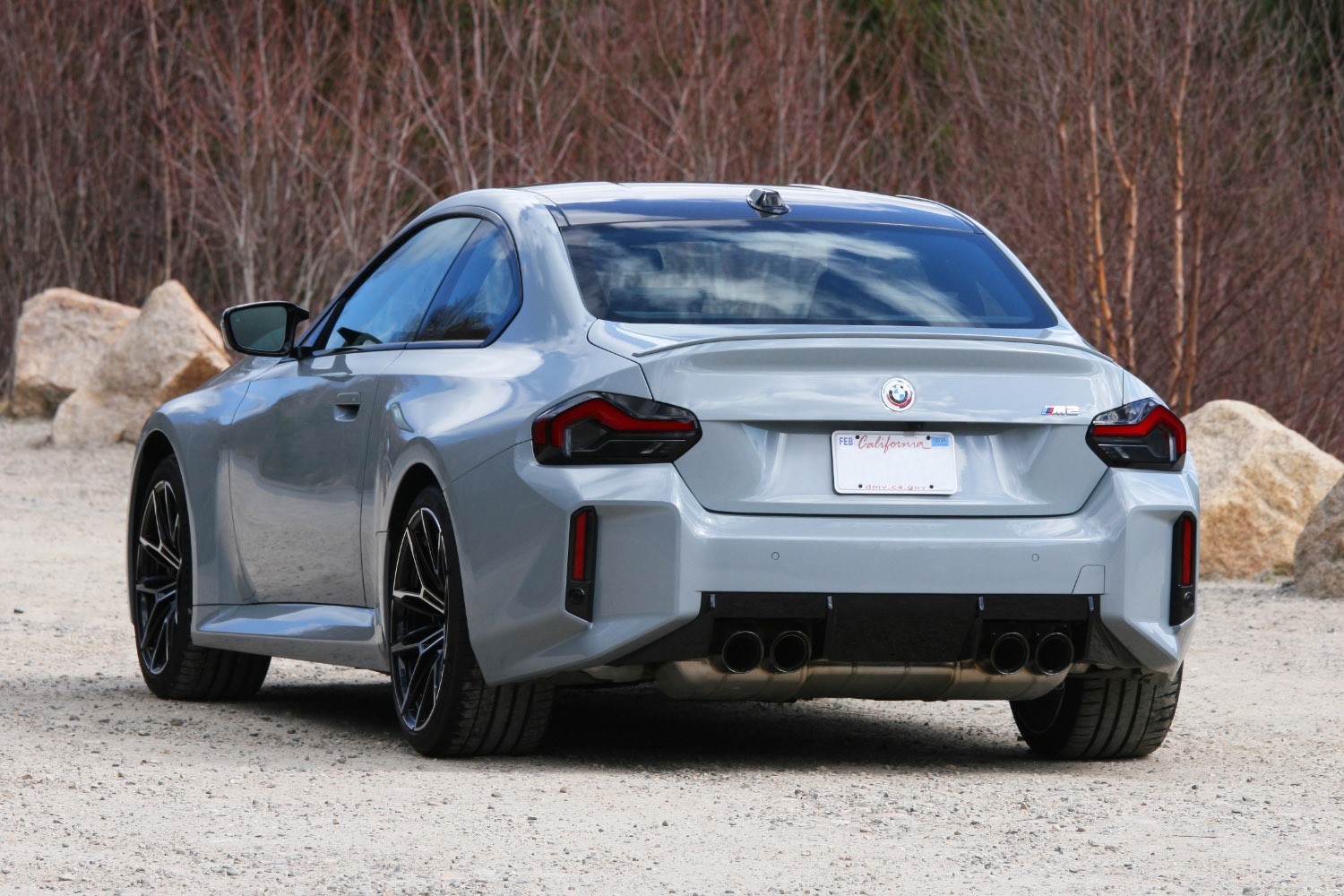 Rear three-quarter shot of a 2023 BMW M2 in Brooklyn Grey Metallic parked on dirt with the New England woods in the background