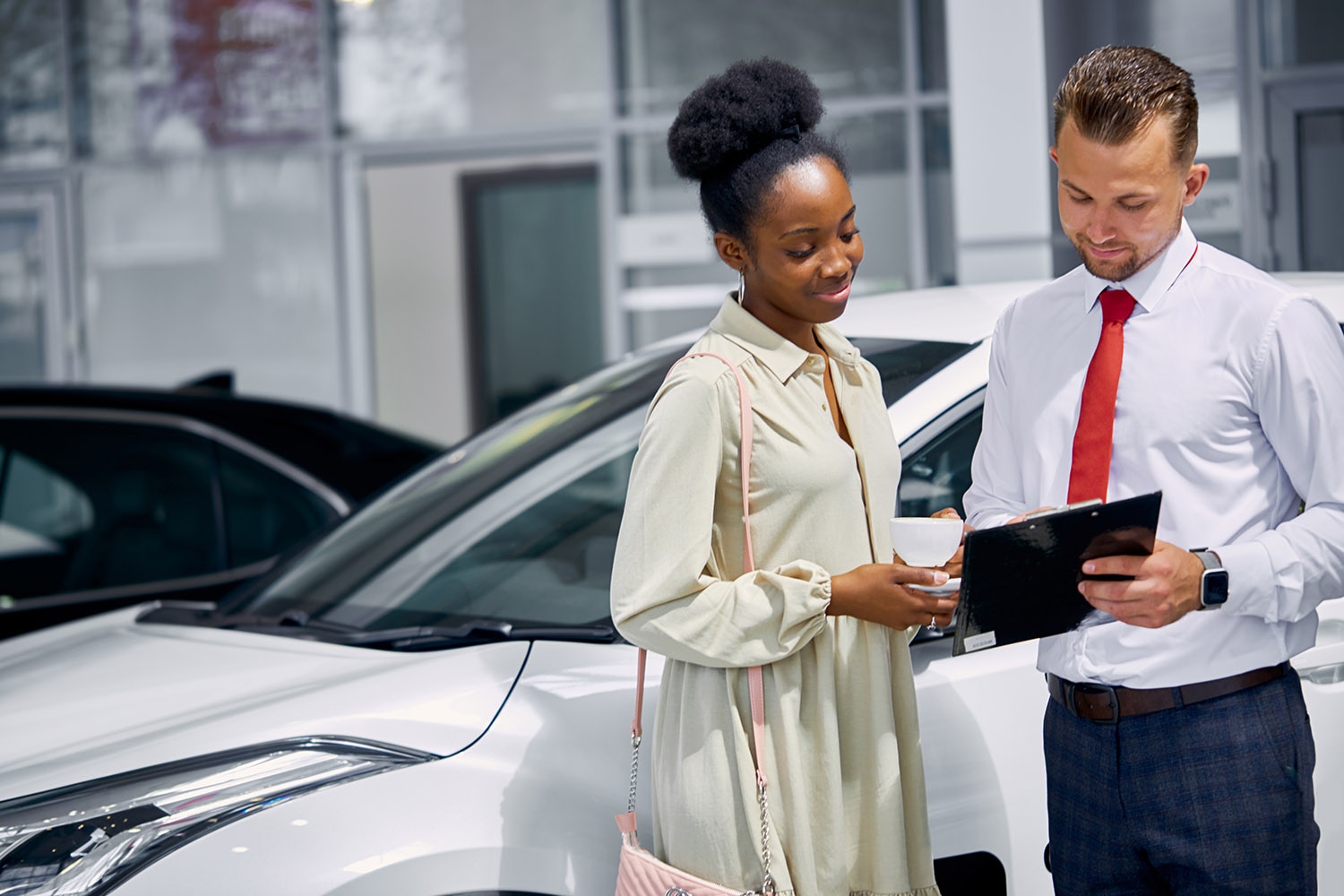 A woman and a man stand in front of a car dealership looking at a clipboard of paperwork