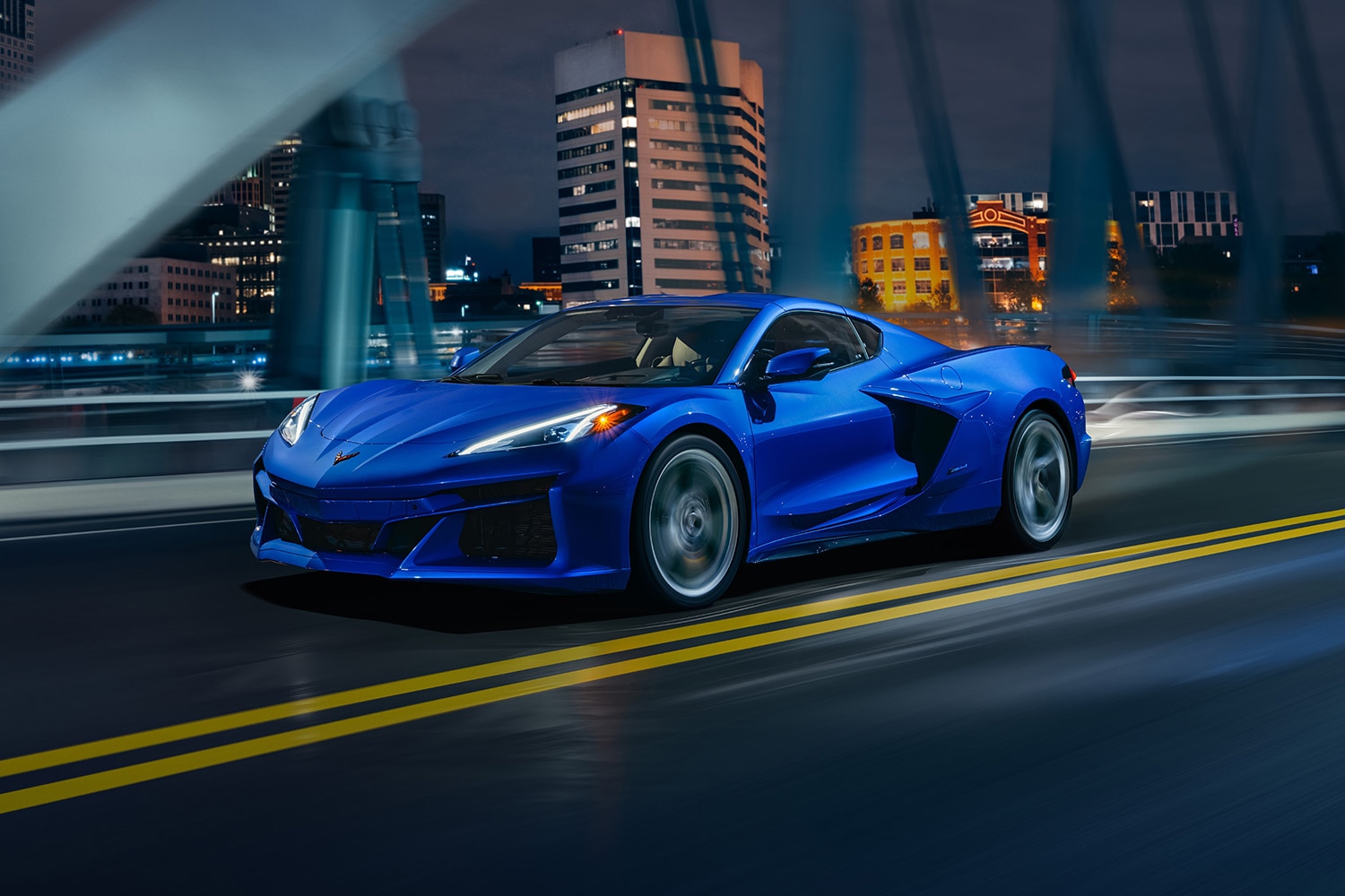 2024 Chevrolet Corvette E-Ray in Riptide Blue driving along a bridge at night with high-rises in the background