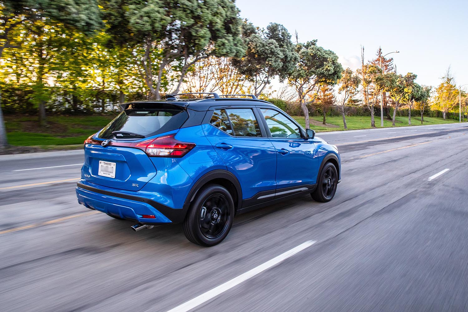 2023 Nissan Kicks in bright blue driving on a paved road beside trees.