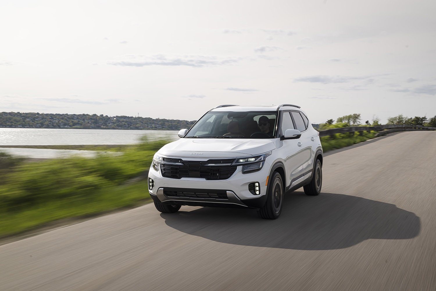 2024 Kia Seltos in white driving on a paved road beside grass and water.