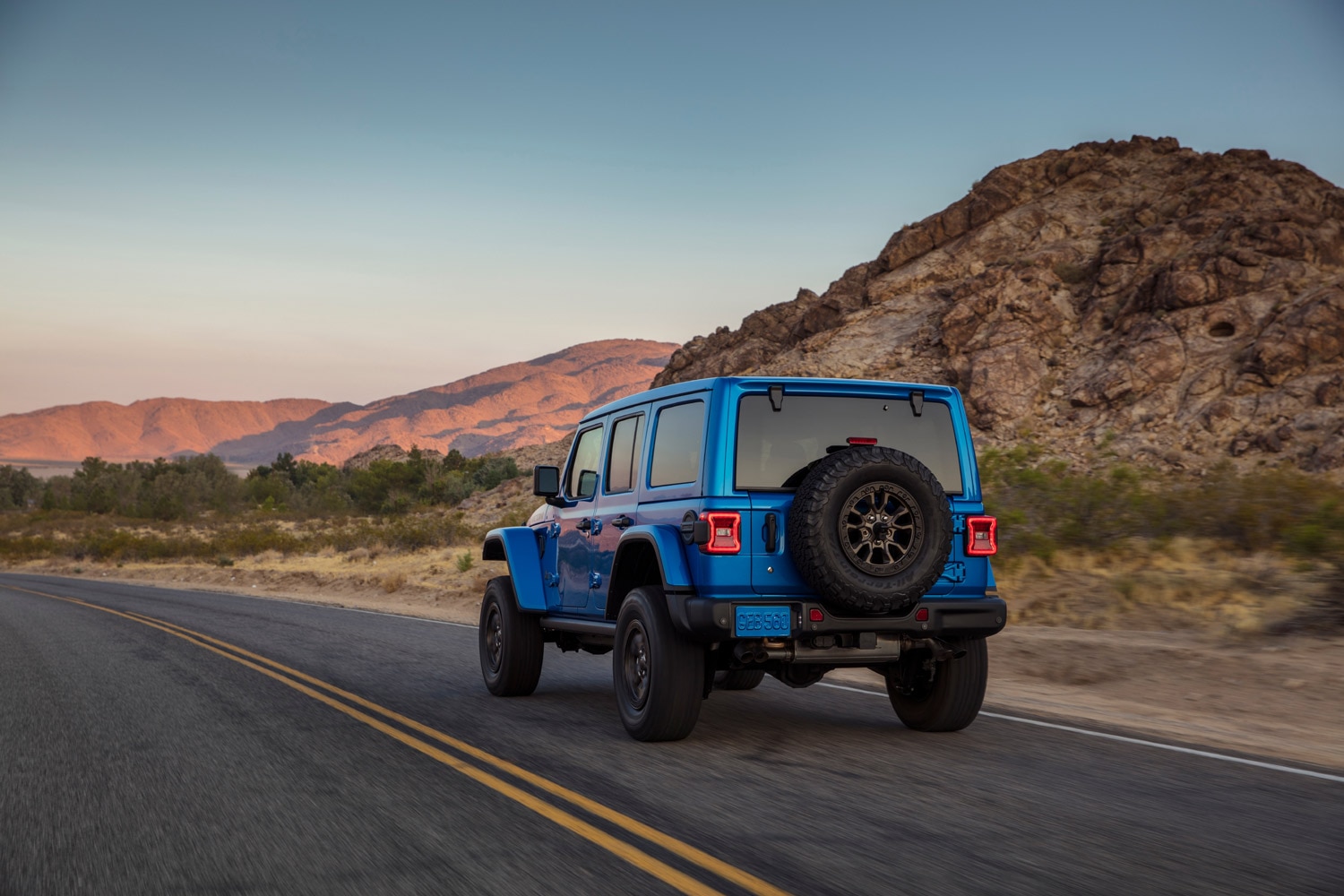 A blue 2023 Jeep Wrangler drives down a desert highway