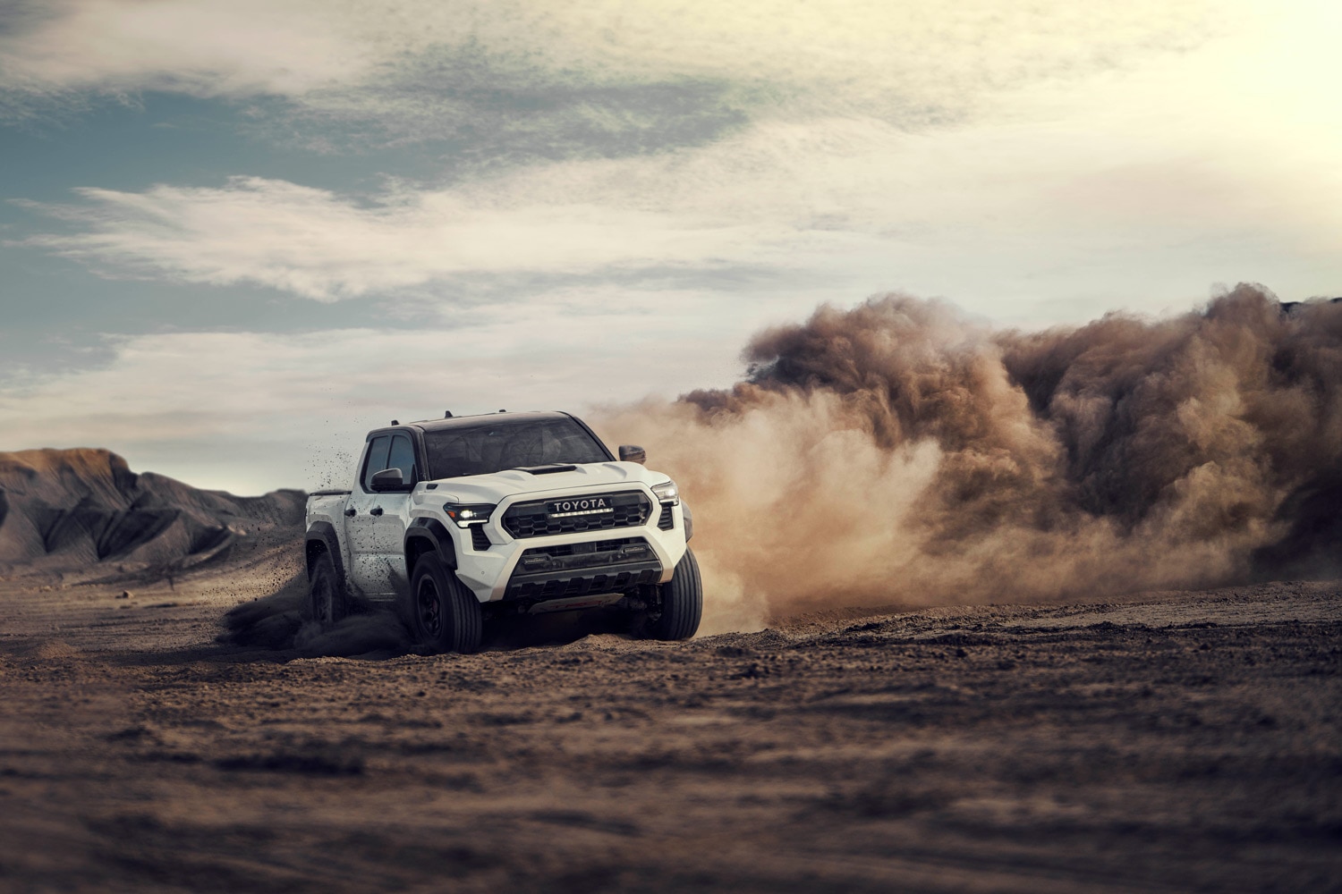 White 2024 Toyota Tacoma TRD Pro driving aggressively on dirt, throwing up dust clouds.