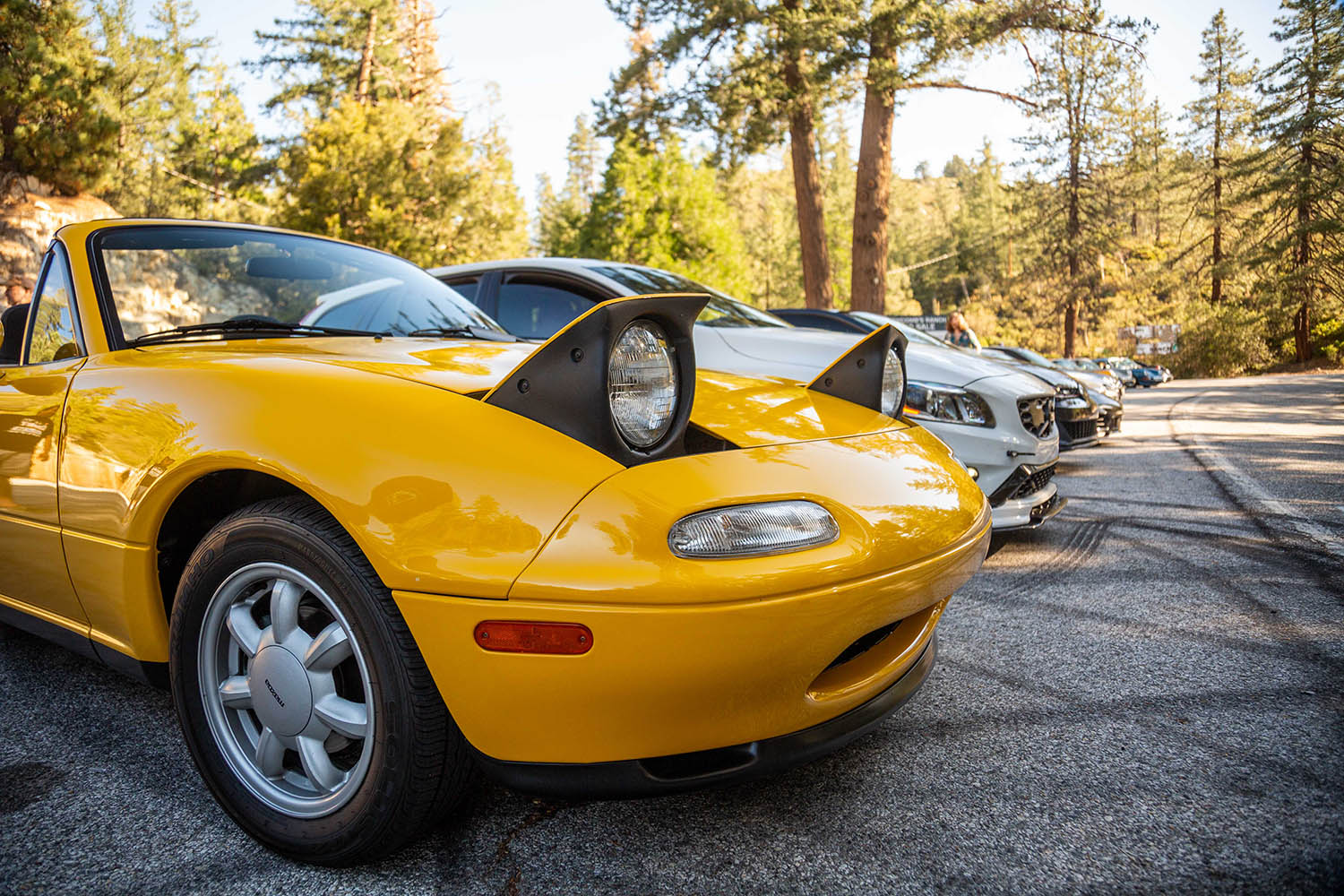 First-generation (NA) Mazda MX-5 Miata in yellow, front-nose view with popup headlights exposed, photographed at Good Vibes Breakfast Club at Newcomb's Ranch on Angeles Crest Highway in Southern California.