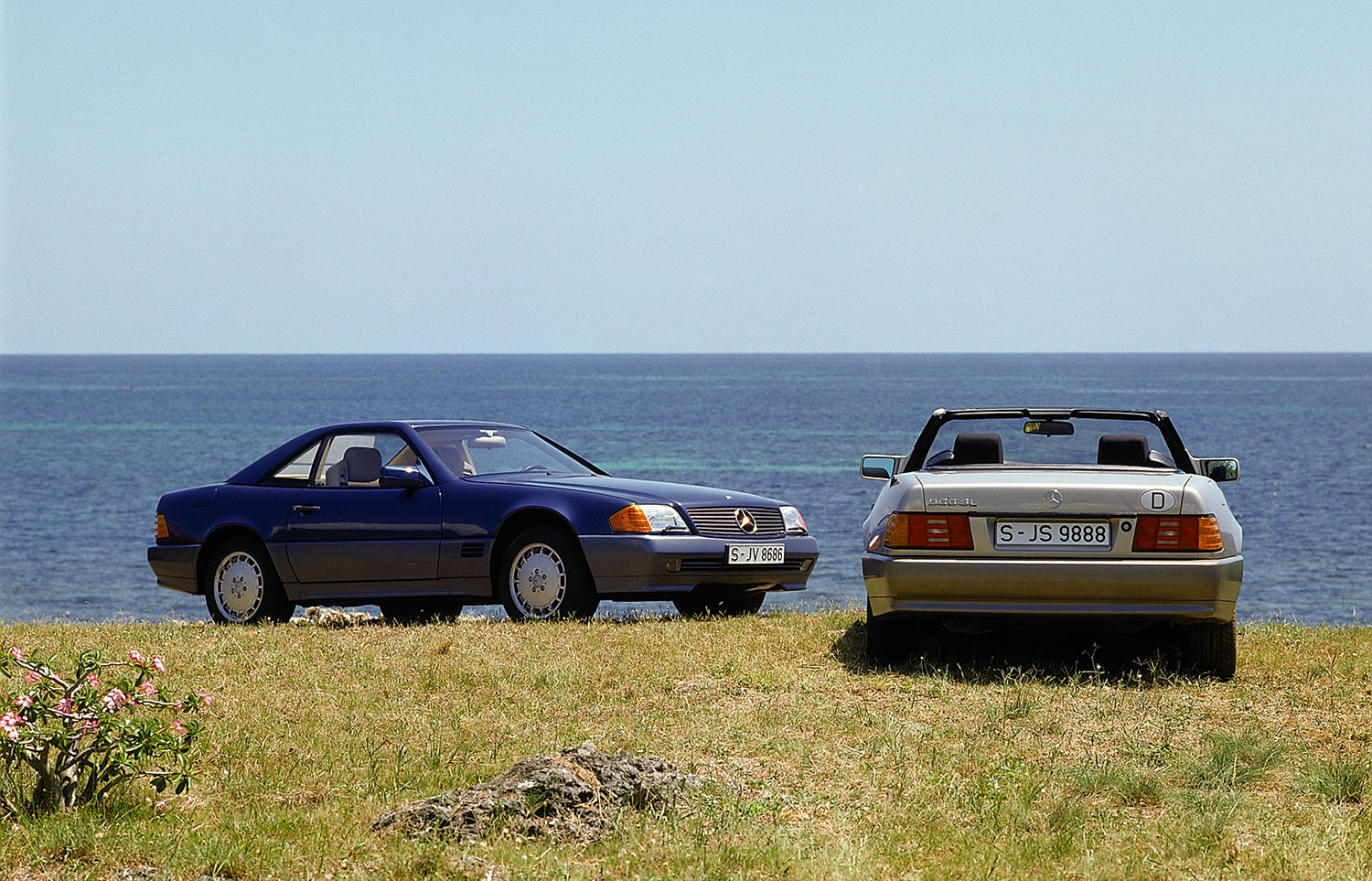 A R129 (1989 to 2001 production) Mercedes-Benz SL in blue with the hard top affixed and one in silver with the soft top stowed, both parked near the sea.