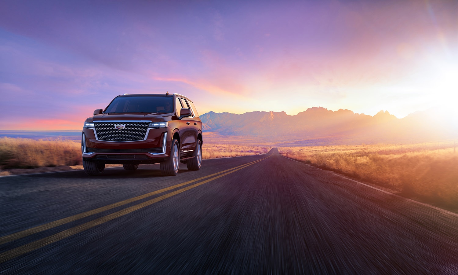 Cadillac Escalade in dark red driving on paved road with sunlight and mountains in the background