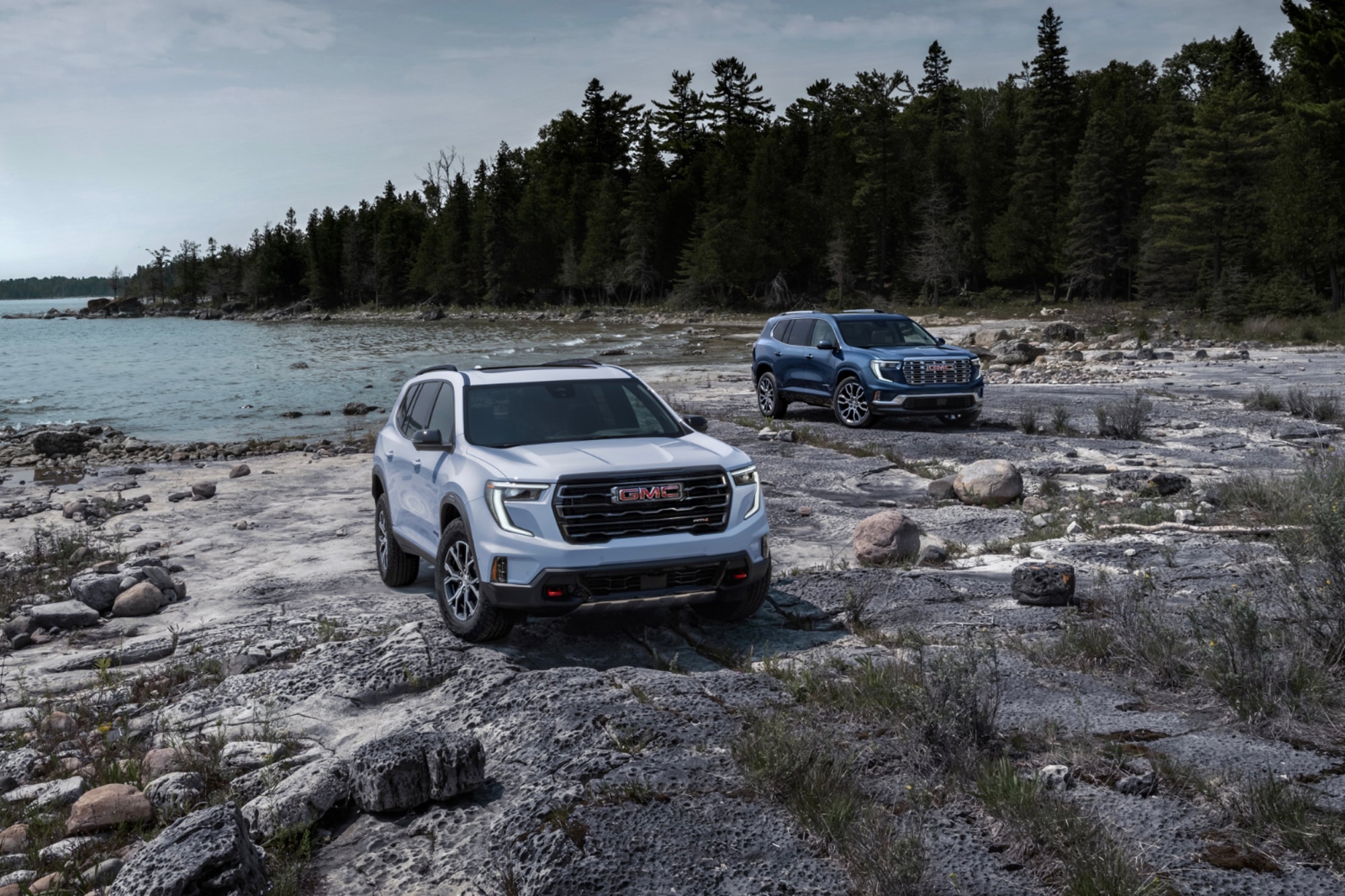 2024 GMC Acadia AT4 in white and GMC Acadia Denali in blue parked by the ocean and forest