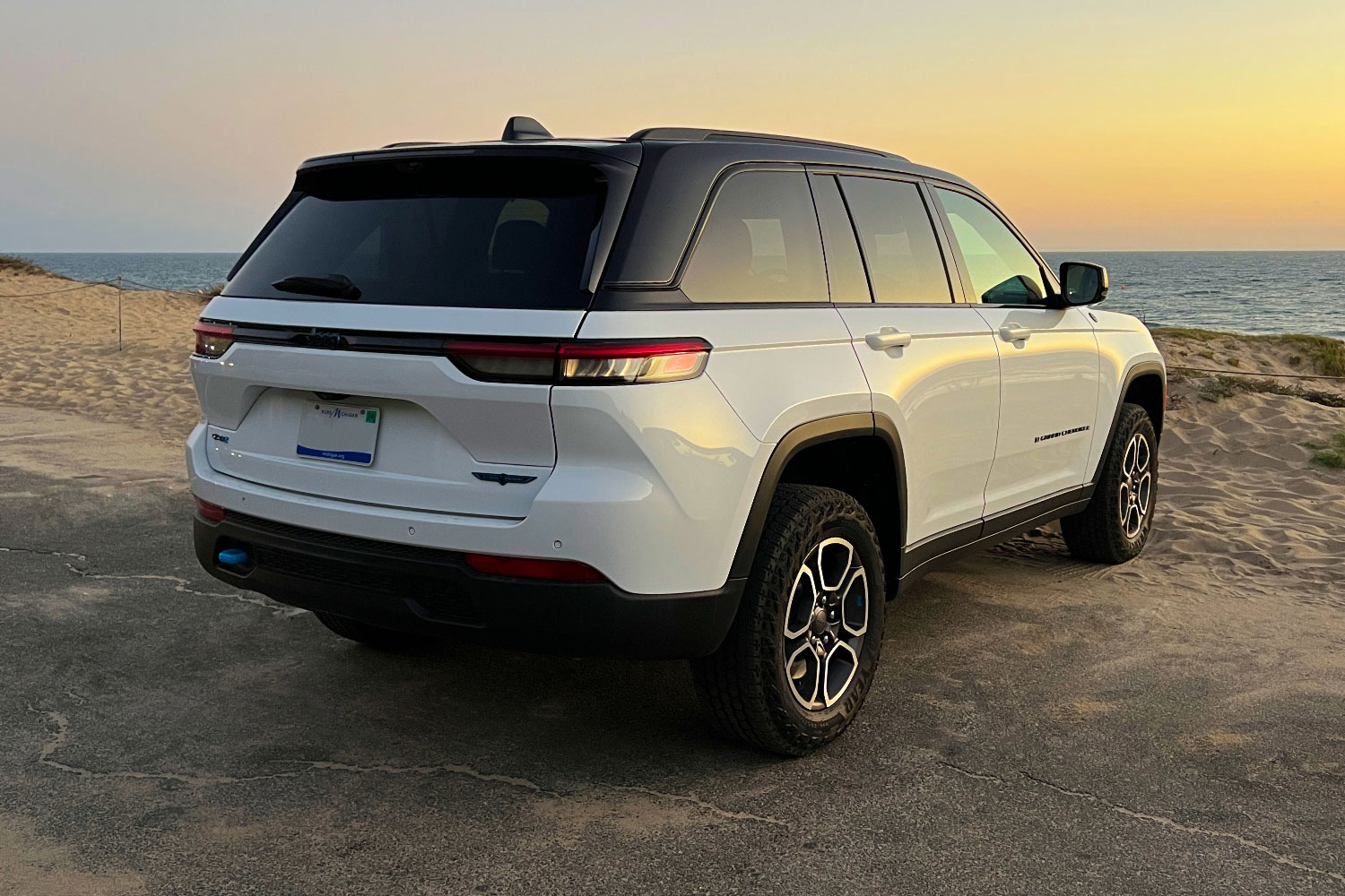 White 2023 Jeep Grand Cherokee 4xe Trailhawk parked by pavement at beach.