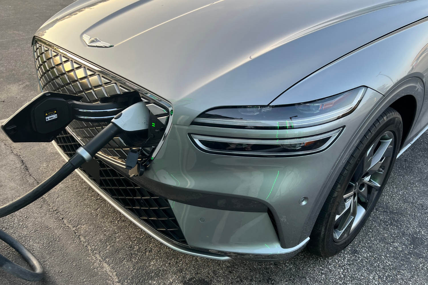 2023 Genesis Electrified GV70 in Savile Silver recharging at a station and showing the grille's hideaway charge port in its open position.