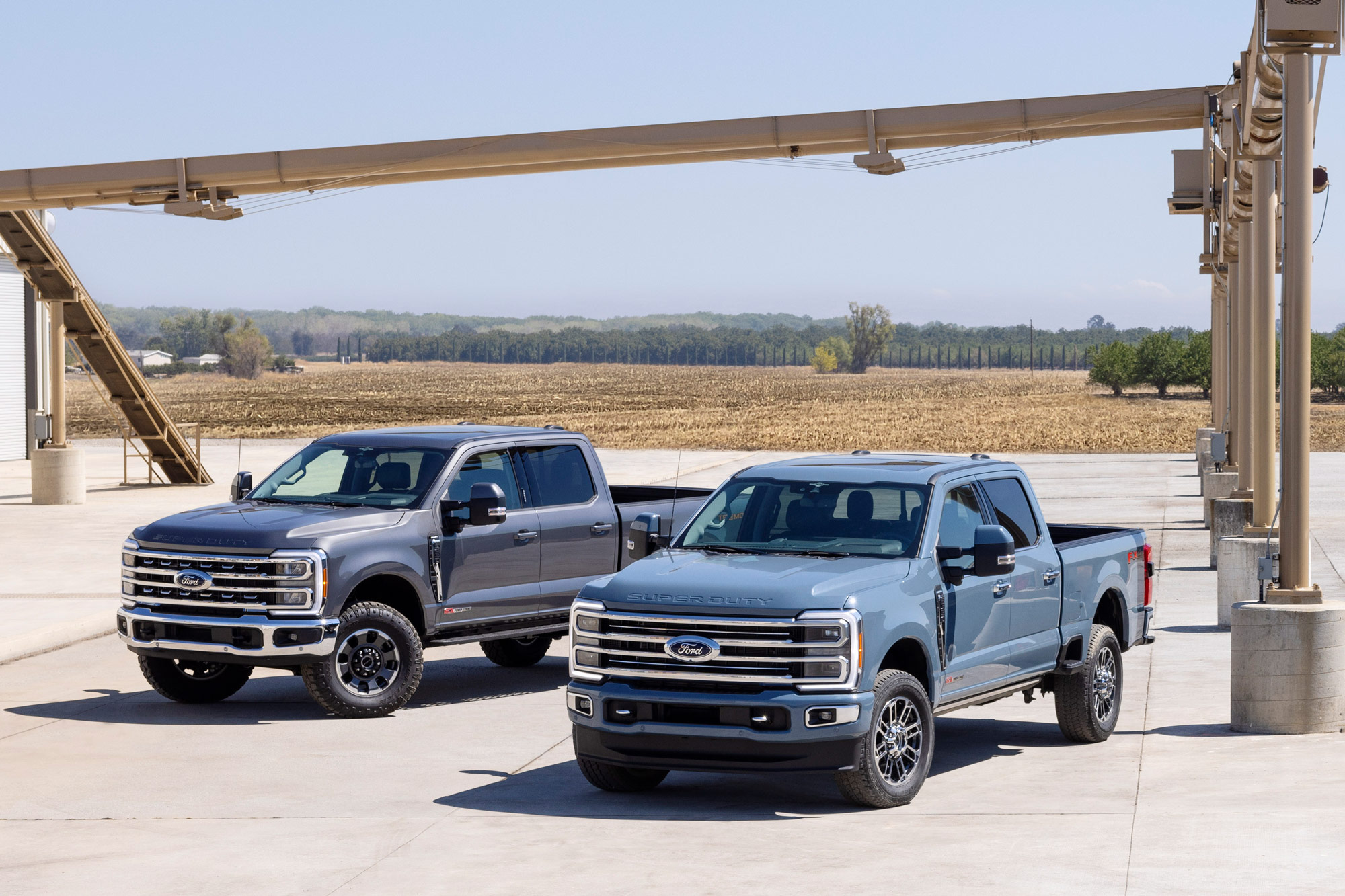 Two 2023 Ford F-Series Super Duty trucks sit parked on a concrete pad by an open field.