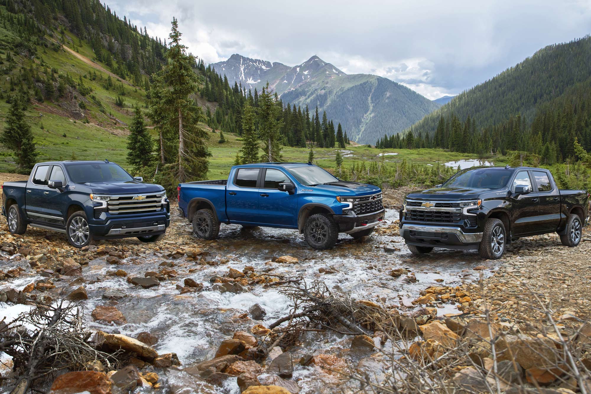 Three 2023 Chevrolet Silverado 1500s sit in and near a small creek in the wilderness.
