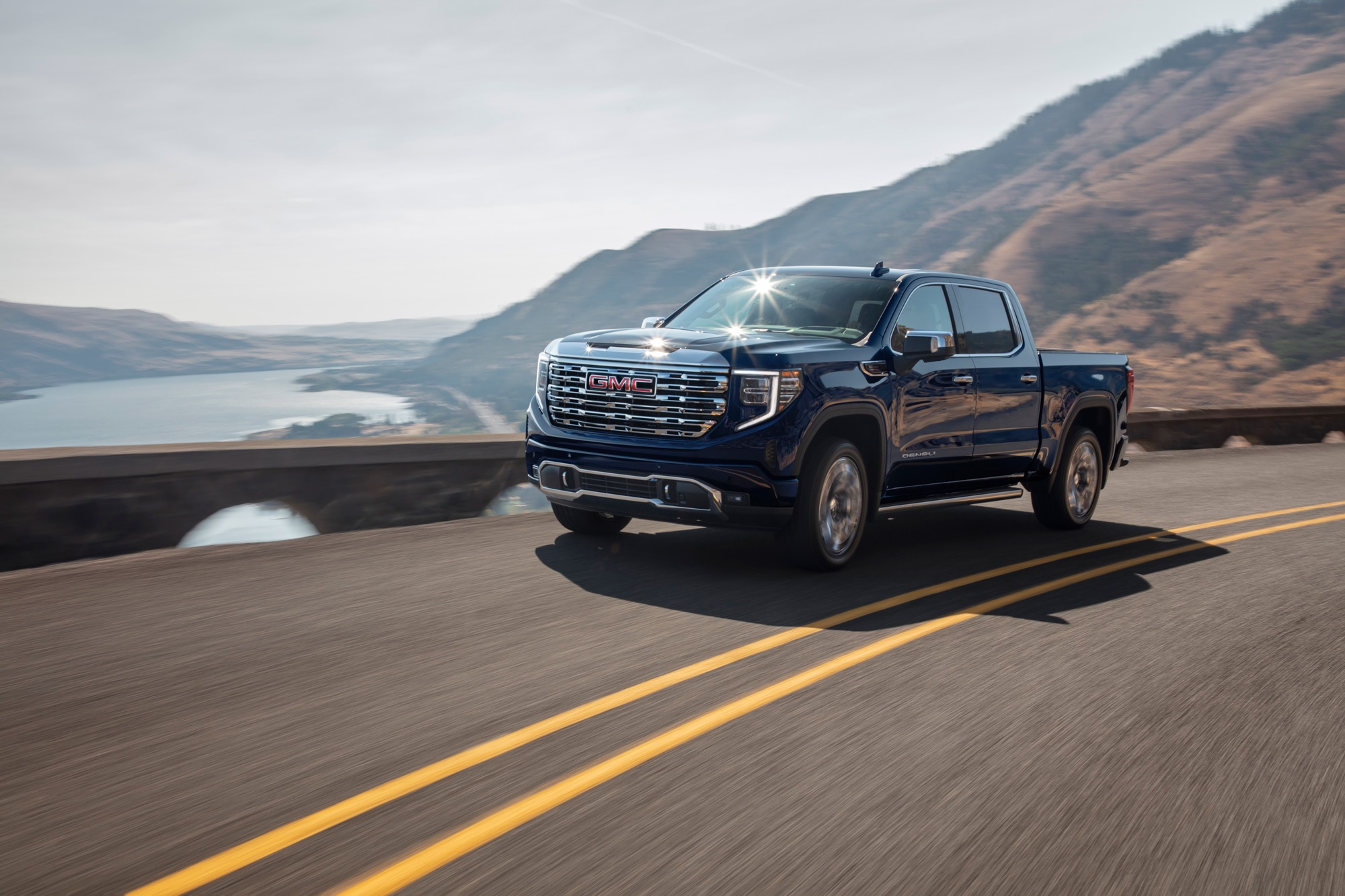 Blue 2023 GMC Sierra 1500 drives along a scenic road overlooking hills and water.