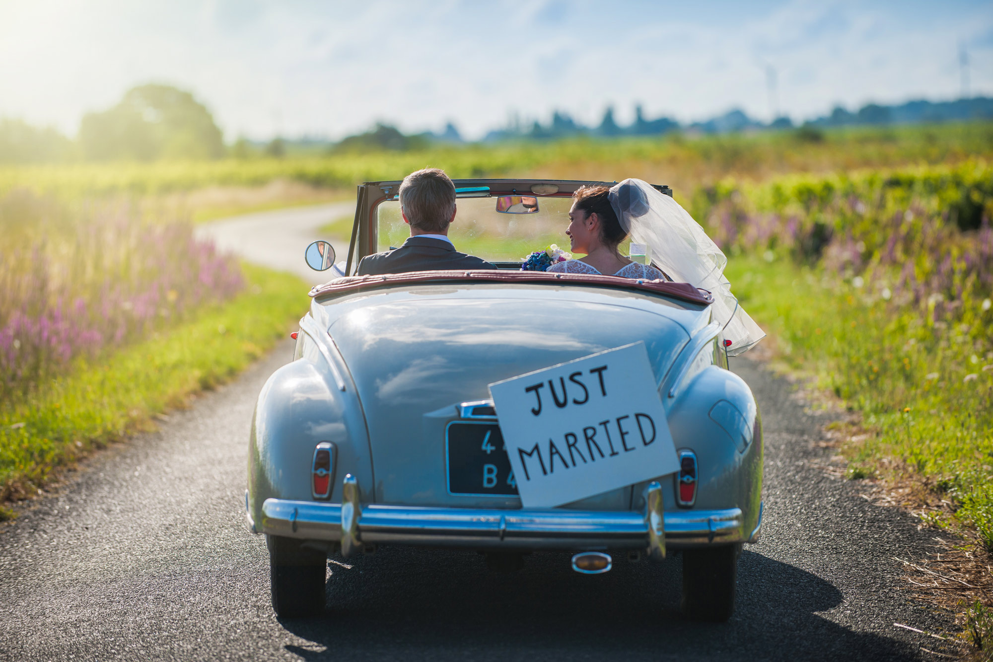 Couple drives down road with just married sign on back of convertible classic car.