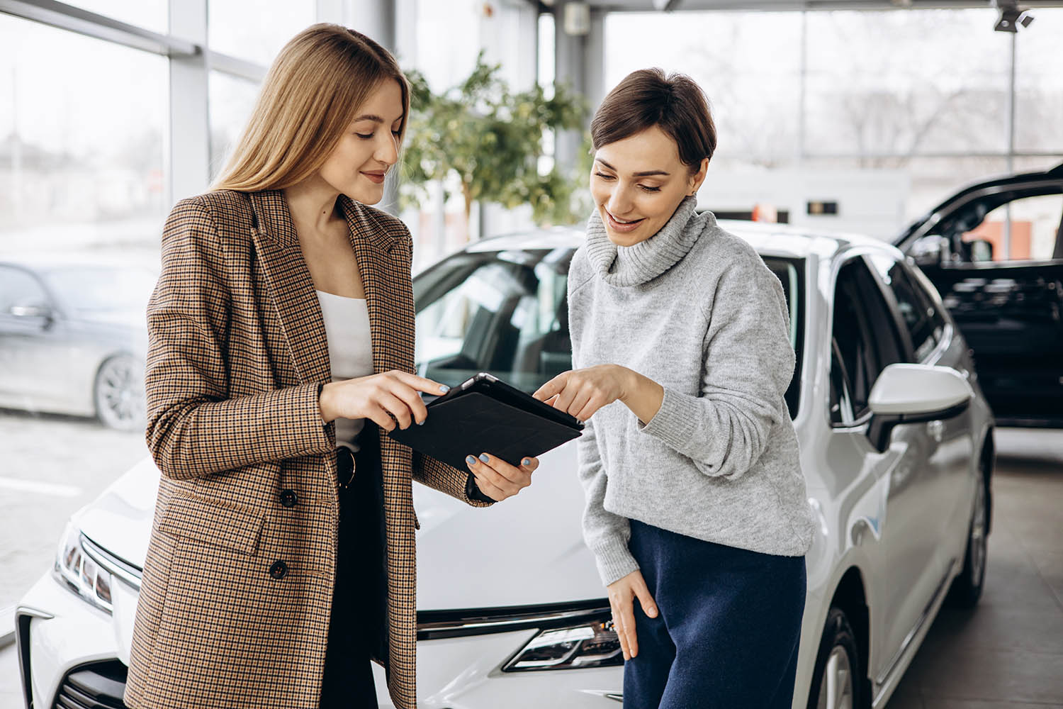 A woman holding out a clipboard to another woman in a car dealership showroom