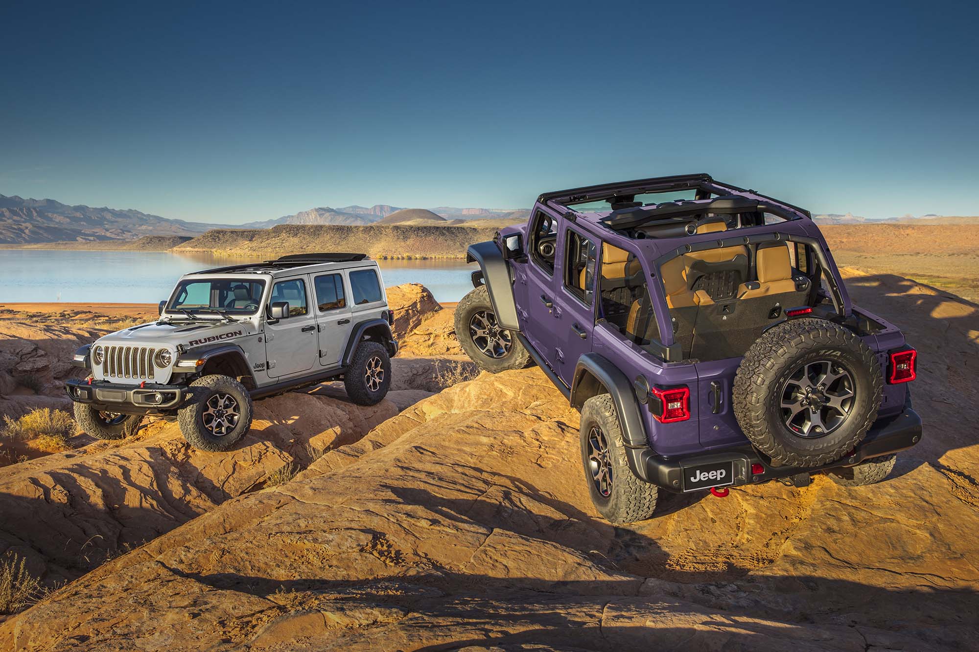 A gray Jeep Wrangler and purple Jeep Wrangler parked on rocks in front of a lake