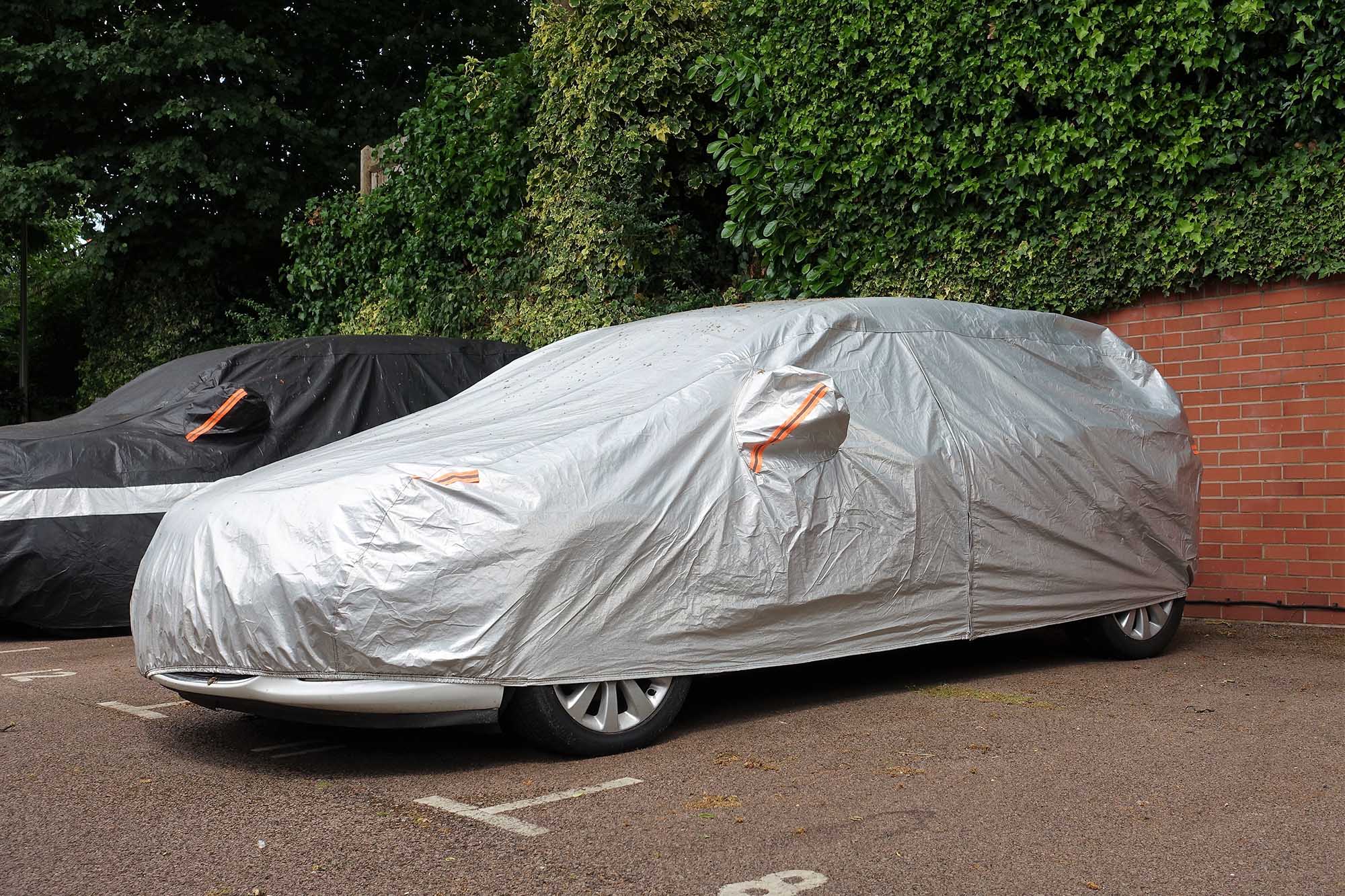 Two cars under vehicle covers parked next to brick wall