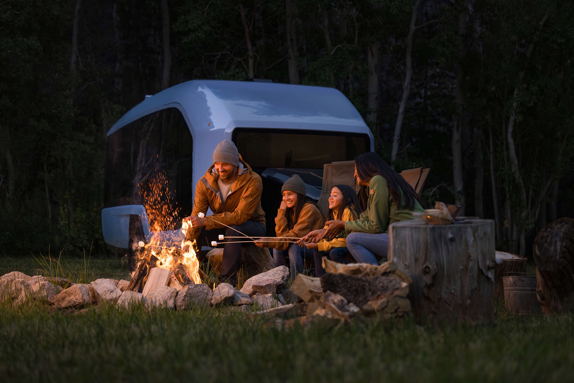 People roast marshmallows over a fire with a Pebble electric camper trailer in the background