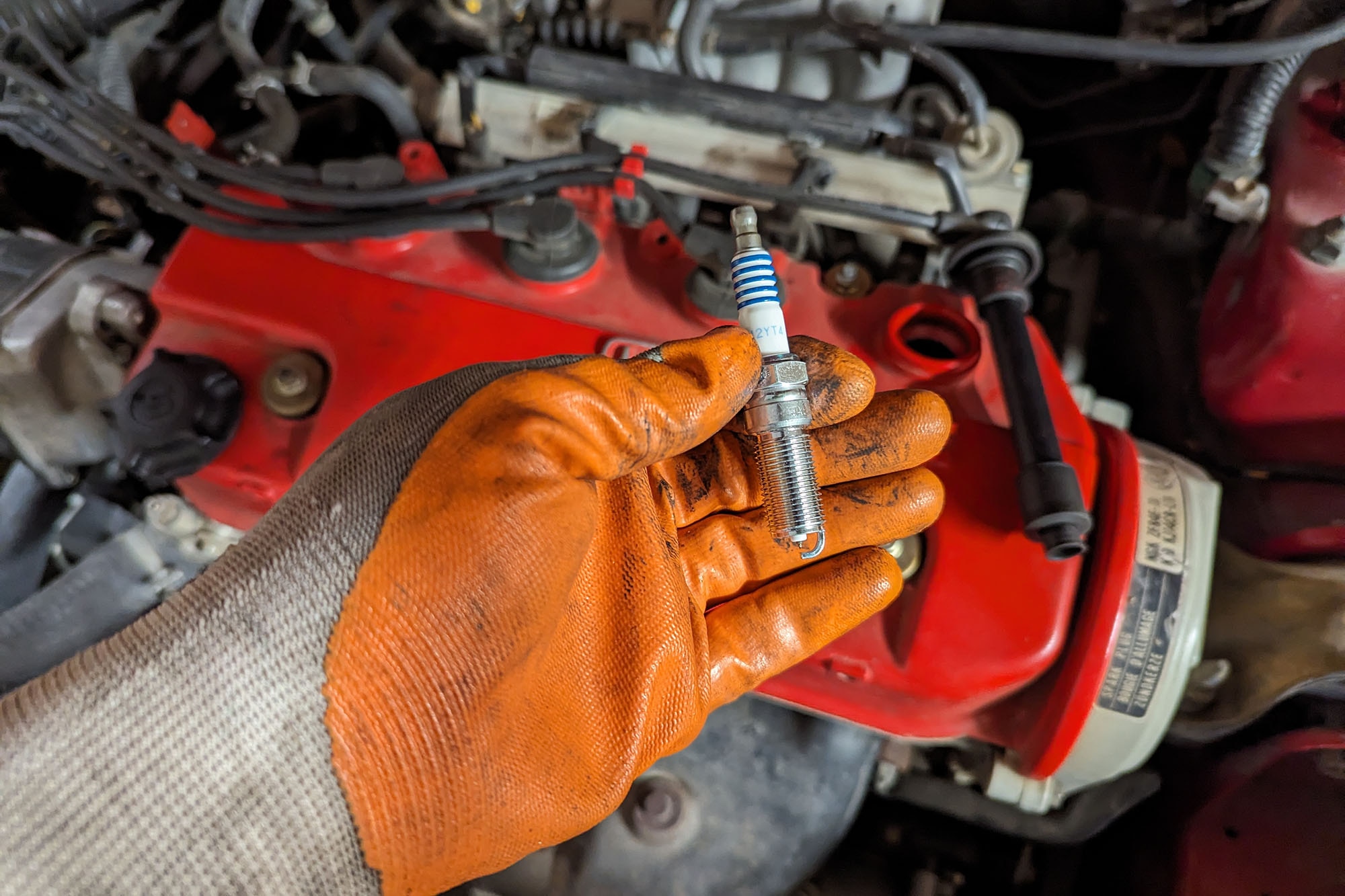 A gloved hand holds a spark plug above an engine bay