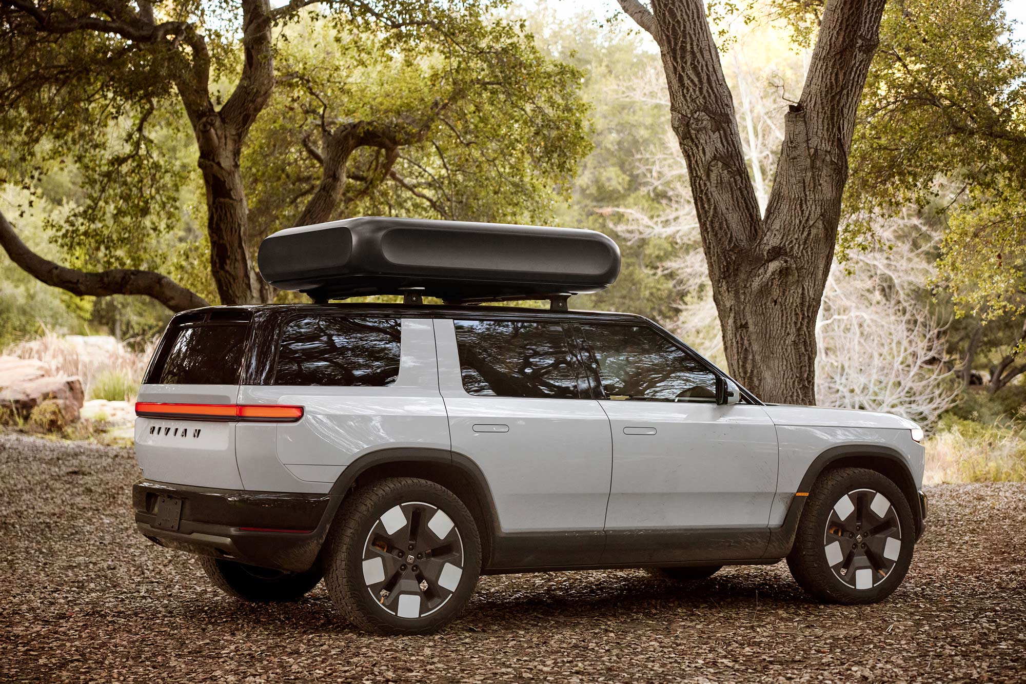 White 2026 Rivian R2 with rooftop box parked under trees.