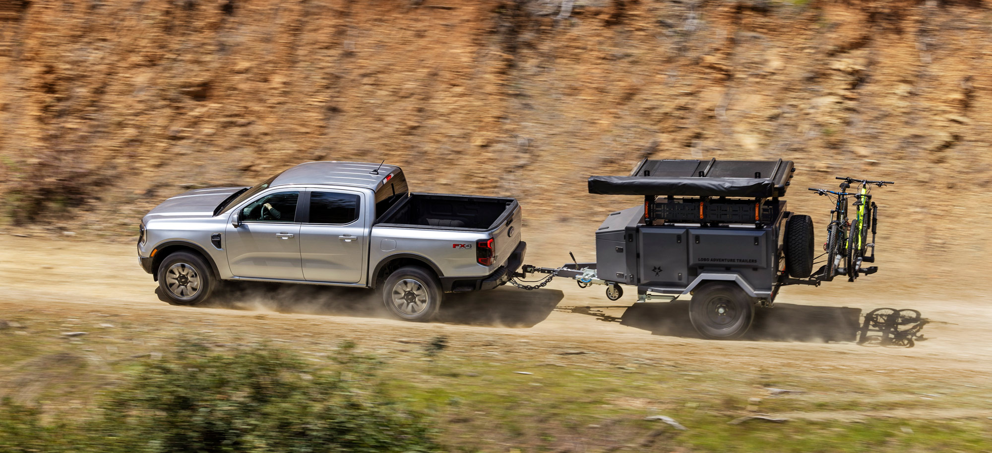Silver 2024 Ford Ranger towing a utility trailer down a dirt road.