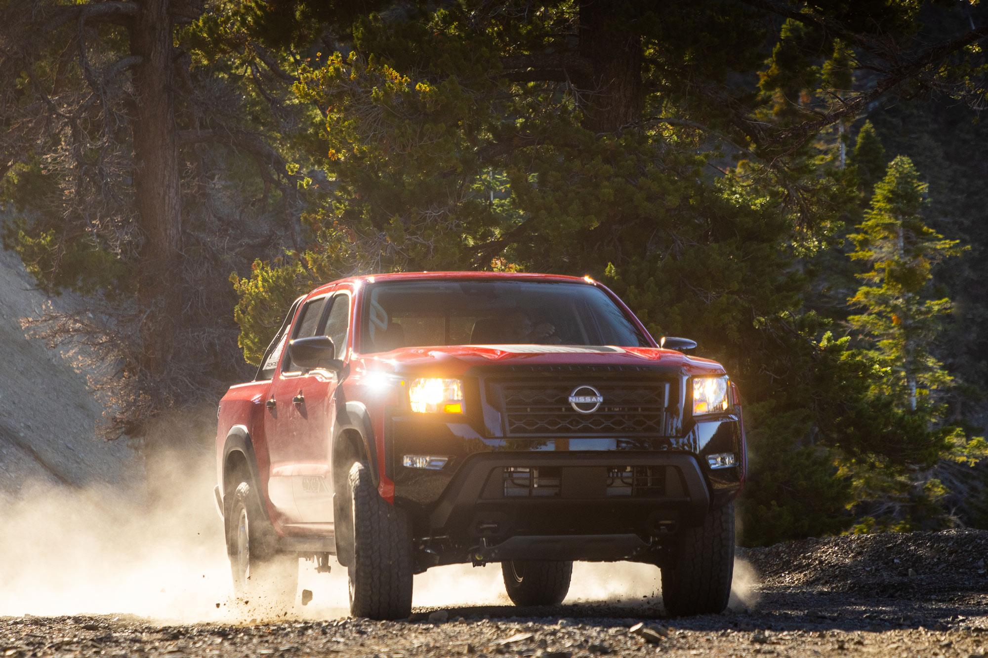 Red 2024 Nissan Frontier drives down forest road throwing up dirt and rocks.