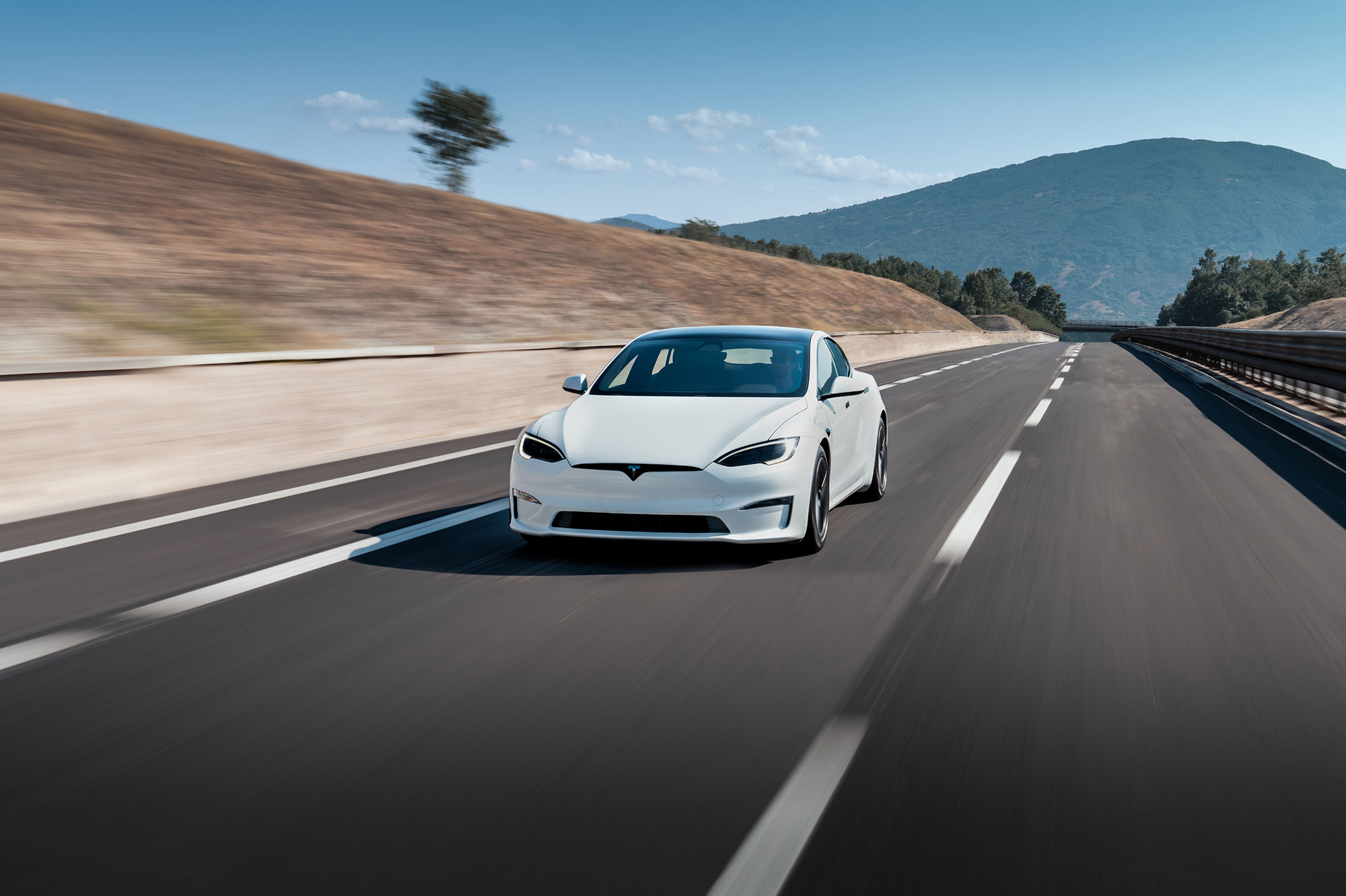 White Tesla Model S driving on paved road with mountains in the background.