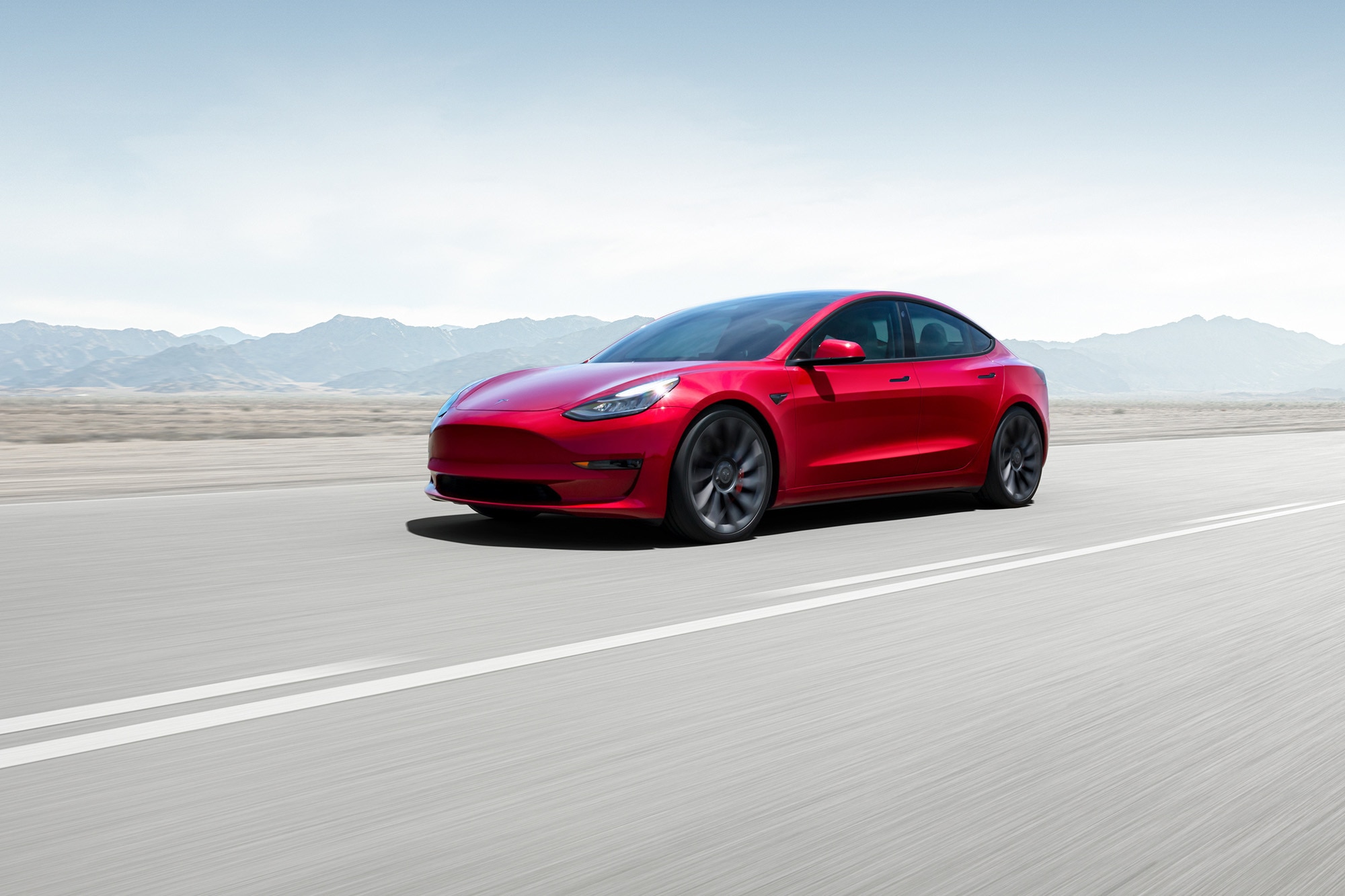 Red Tesla Model 3 driving on paved road with mountains in background.