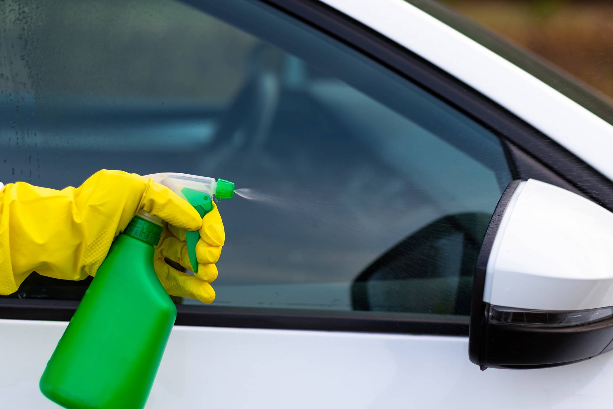 A hand in a yellow glove sprays liquid onto a car from a green bottle.
