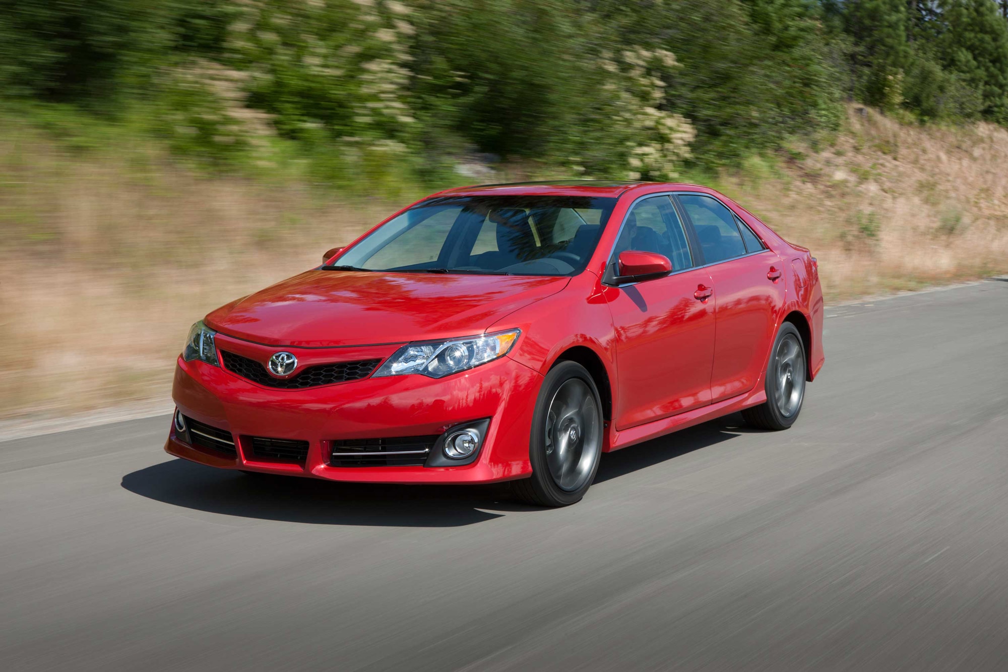 Red 2014 Toyota Camry driving on a road by trees.