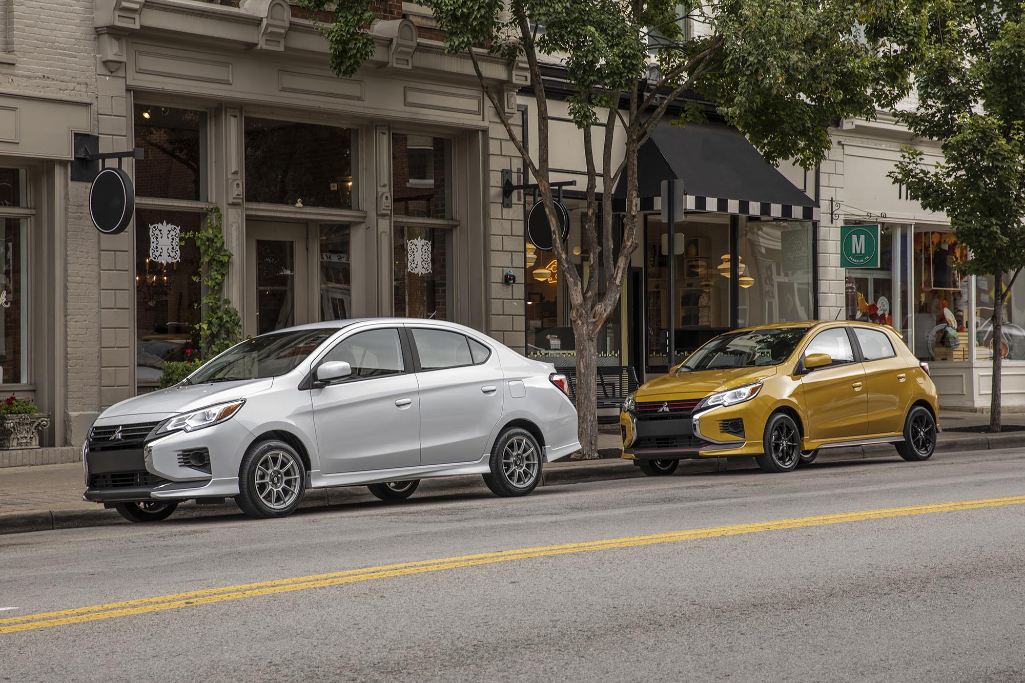 Two Mitsubishi Mirages in silver and yellow parked in front of stores.