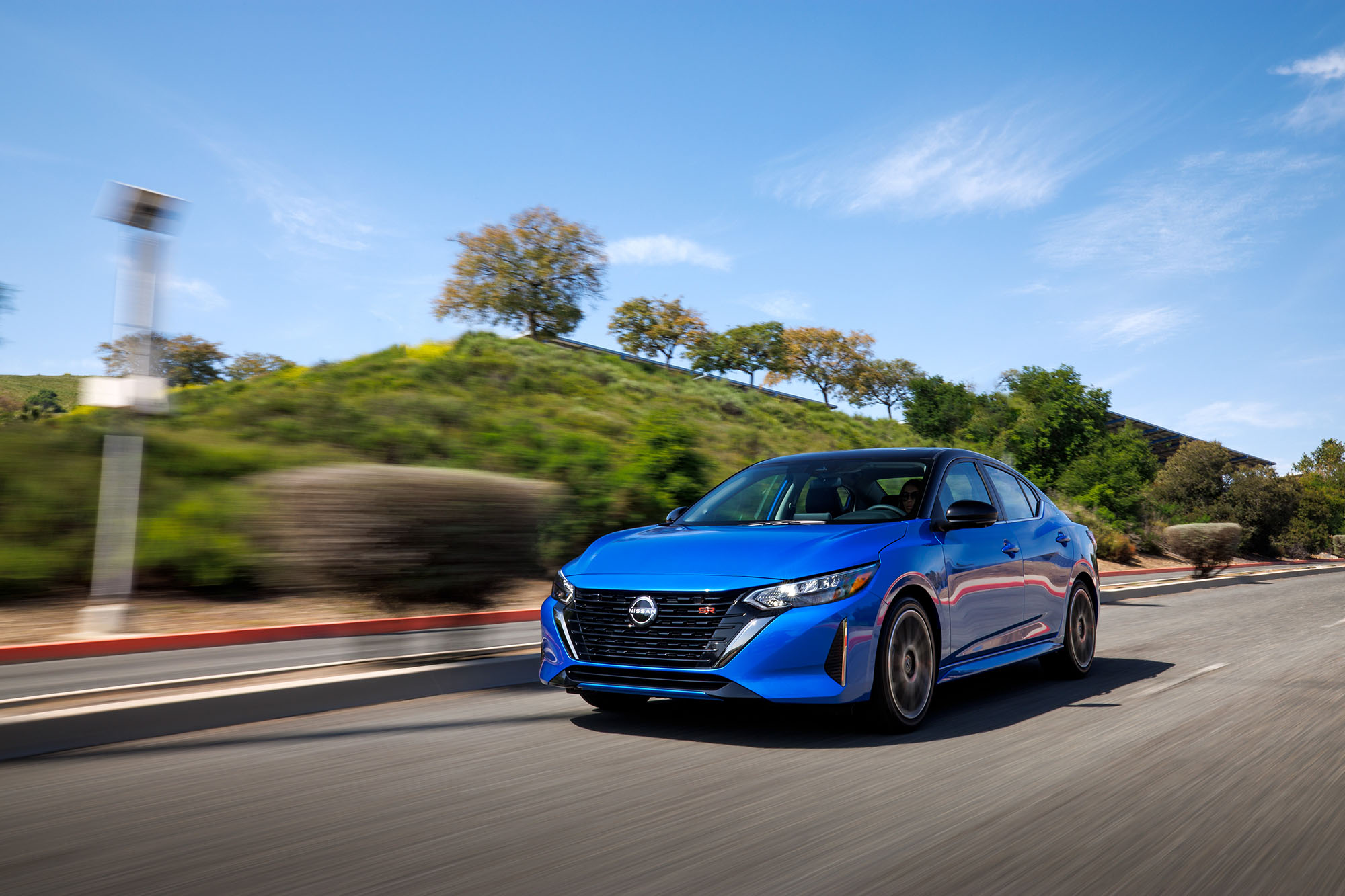 Blue Nissan Sentra driving on road past green hill.